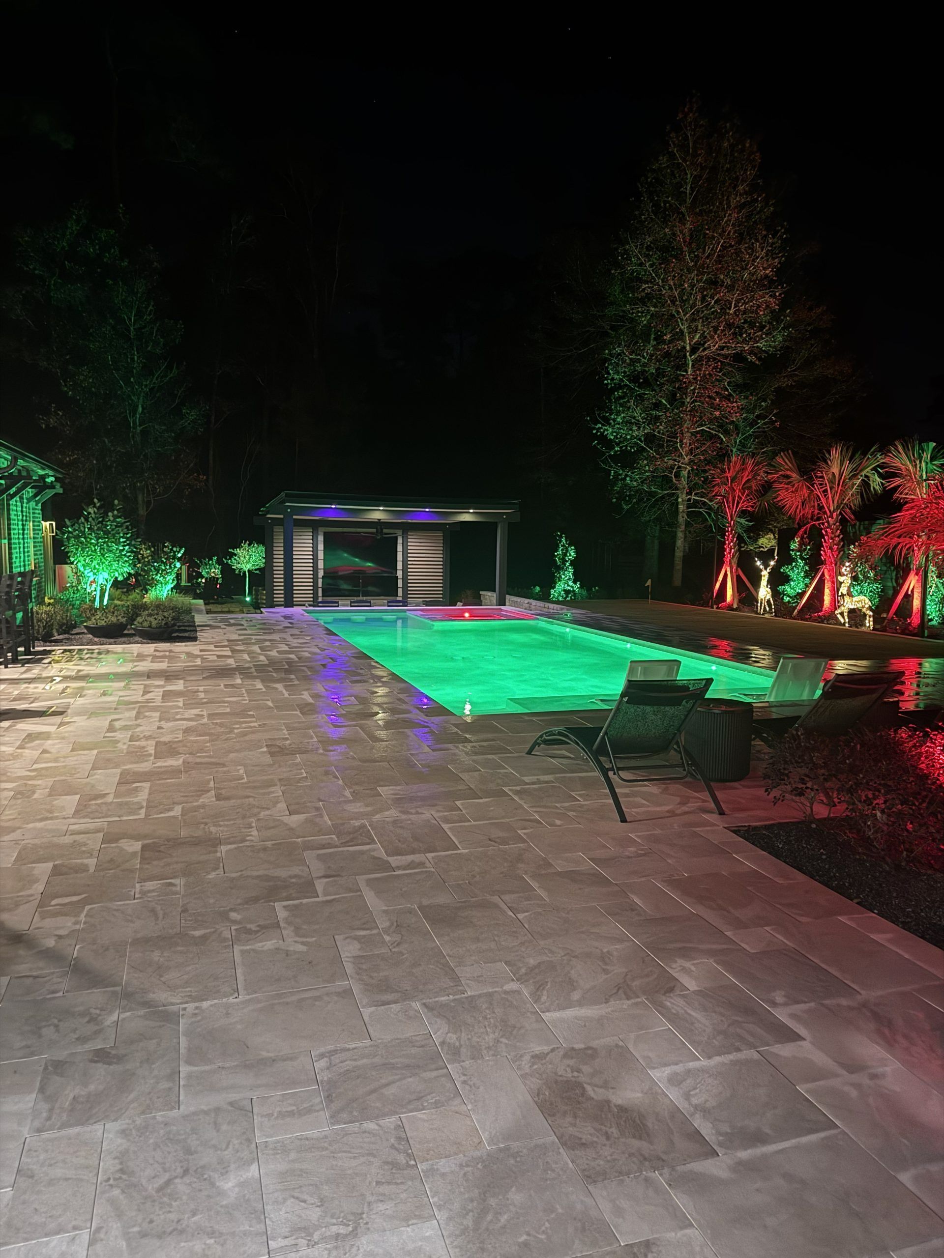 Nighttime poolside patio with glowing green water, stone paving, and colorful landscape lighting