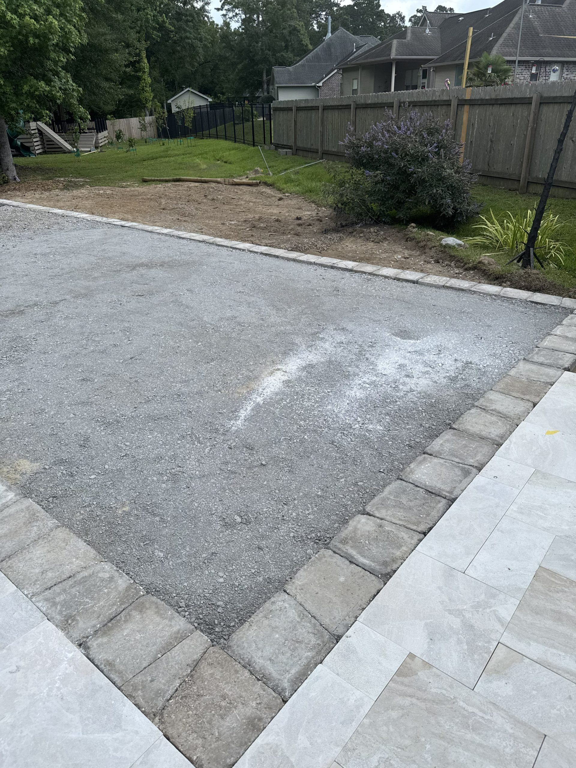 Gravel driveway beside a fenced yard and grass, with gray pavers bordering the edge.