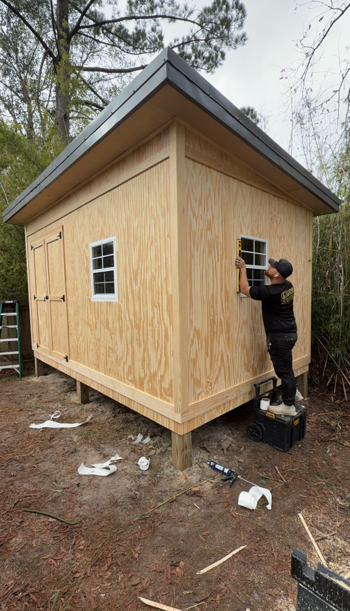 Small unfinished wooden cabin with a dark roof, shown outdoors while a person works on the side.
