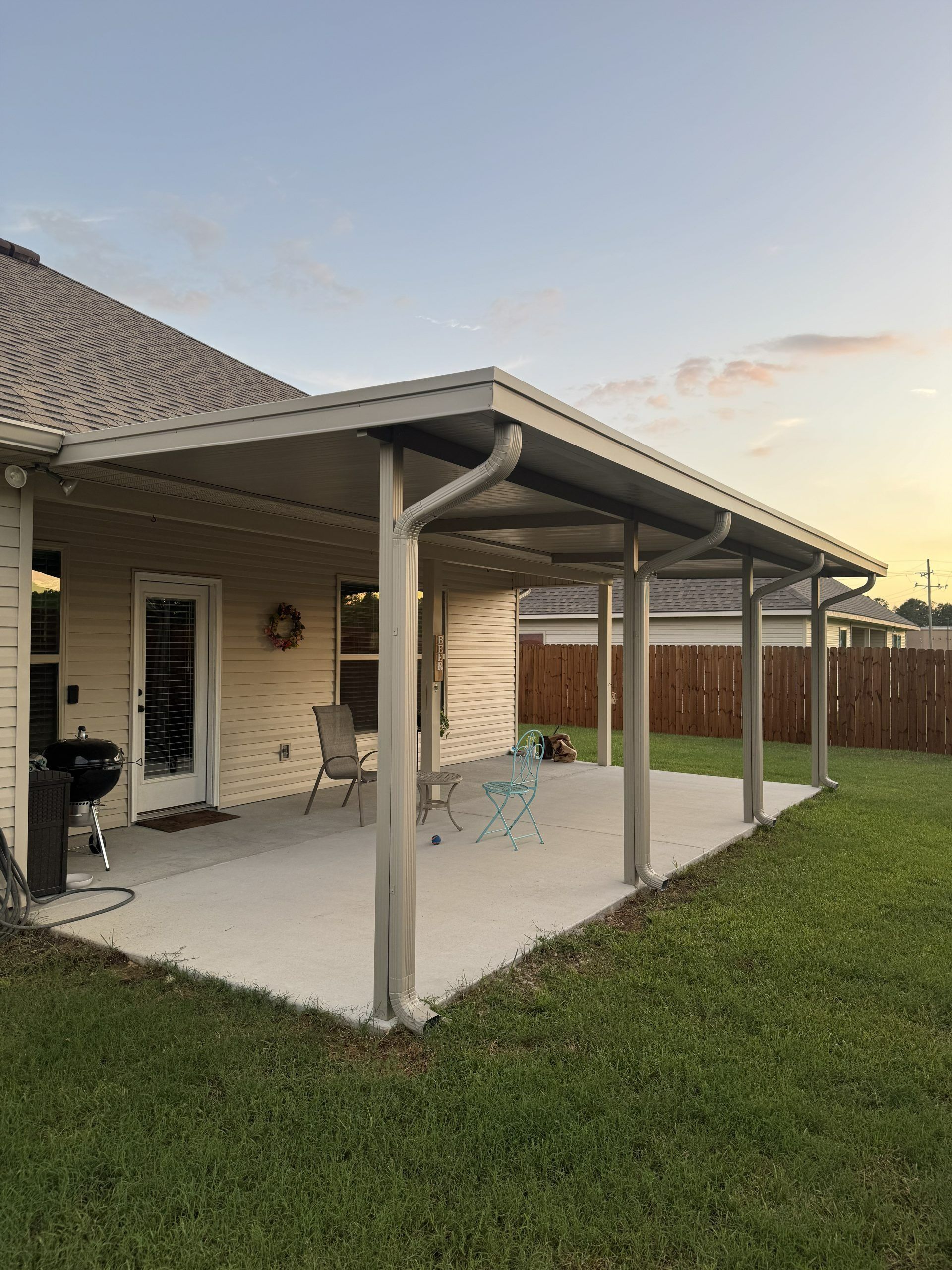Covered patio beside a beige house with gray posts, a grill, and a grassy backyard at sunset