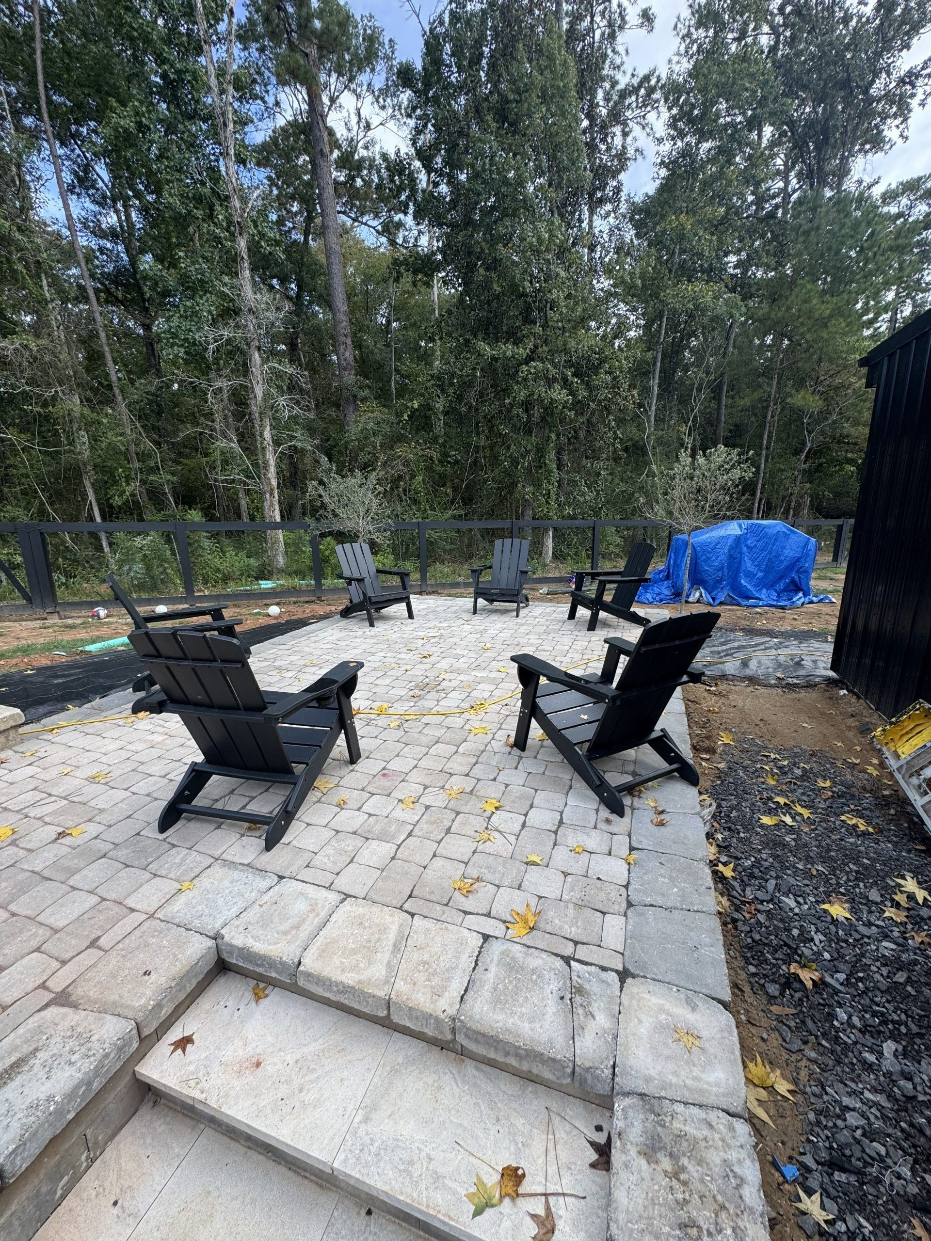 Back patio with two black chairs, stone paving, trees, and a blue tarp-covered item in the background