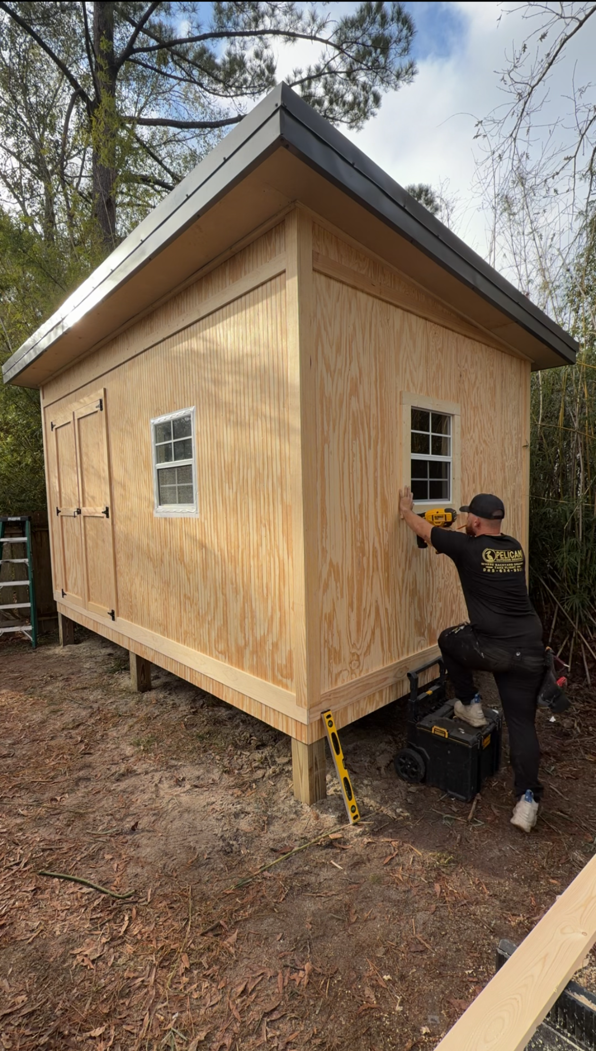 Person using tools to work on the side of a small wooden shed in a wooded yard.