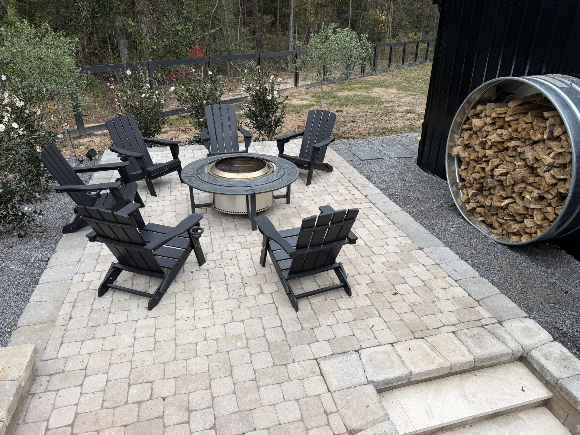 Patio with four black chairs around a fire pit beside stacked firewood and steps