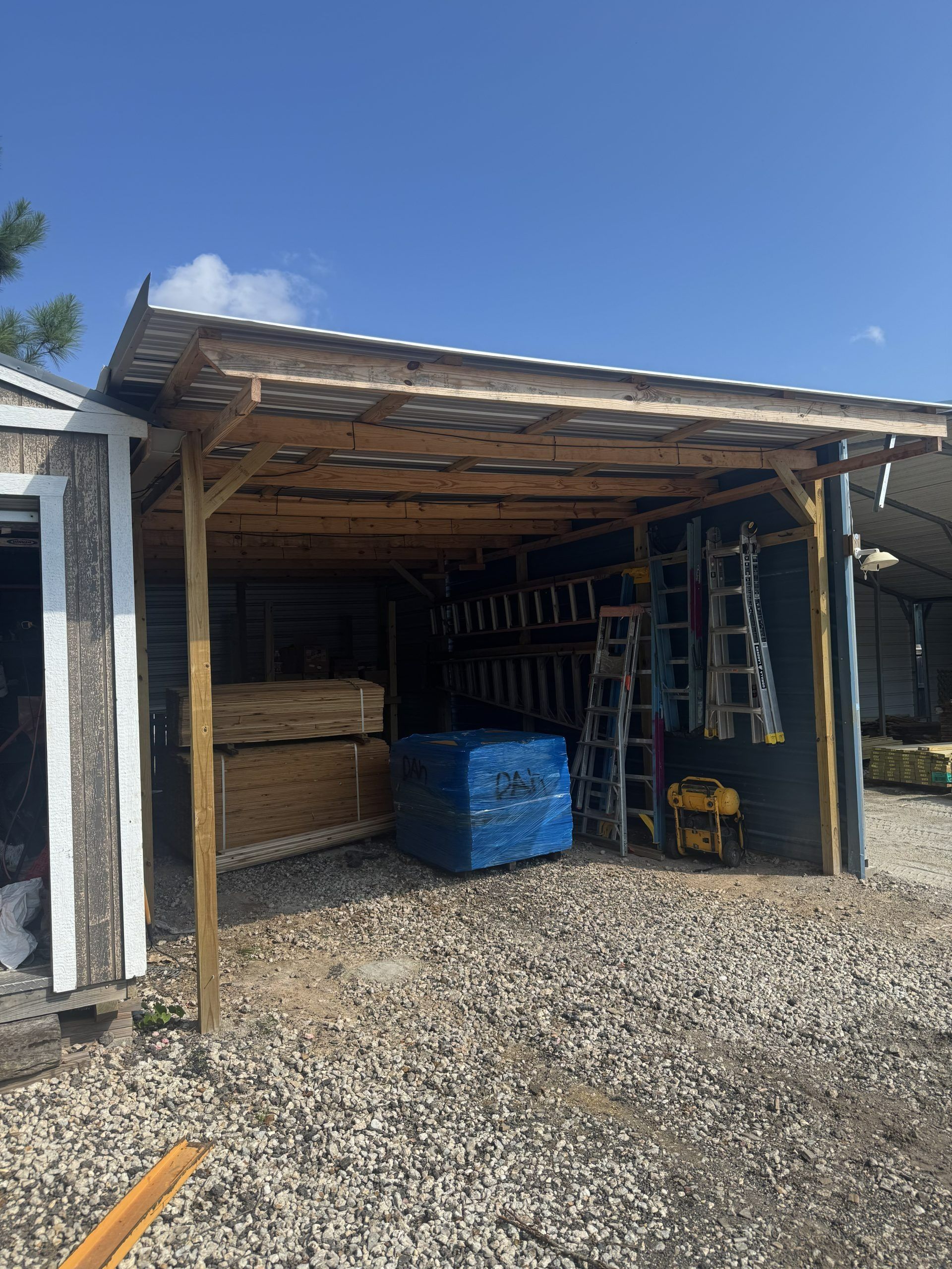 Open-sided shed with stacked wood, a blue bin, and a ladder on a gravel driveway under a blue sky
