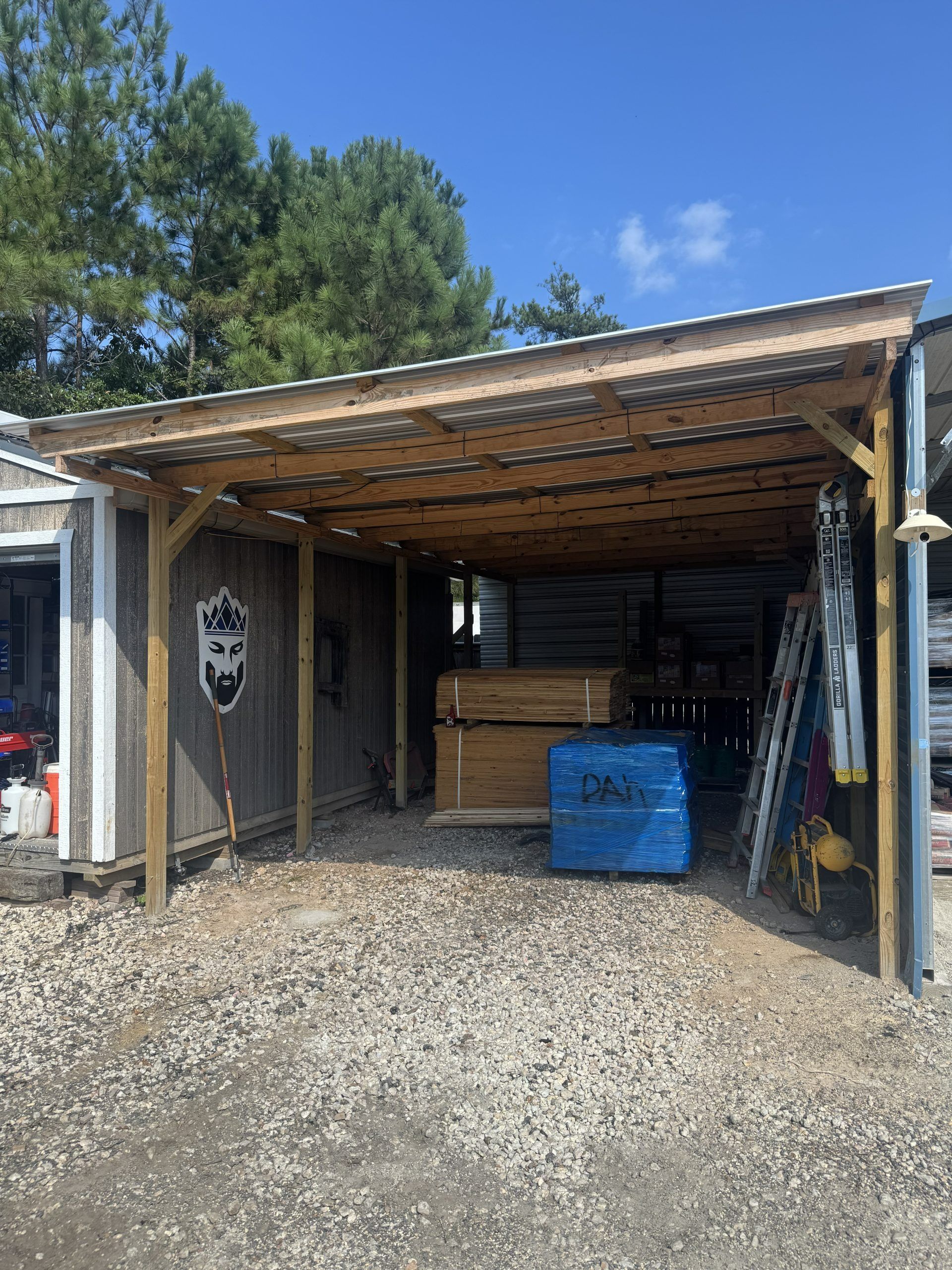 Open-sided wooden shed with tools, a blue bin, and gravel driveway under a clear sky.