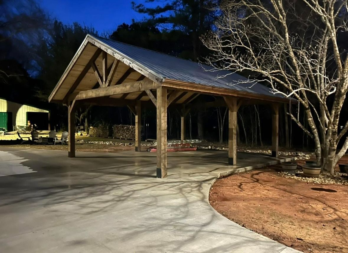 Wooden pavilion with a gabled roof beside a paved driveway at dusk, surrounded by trees.