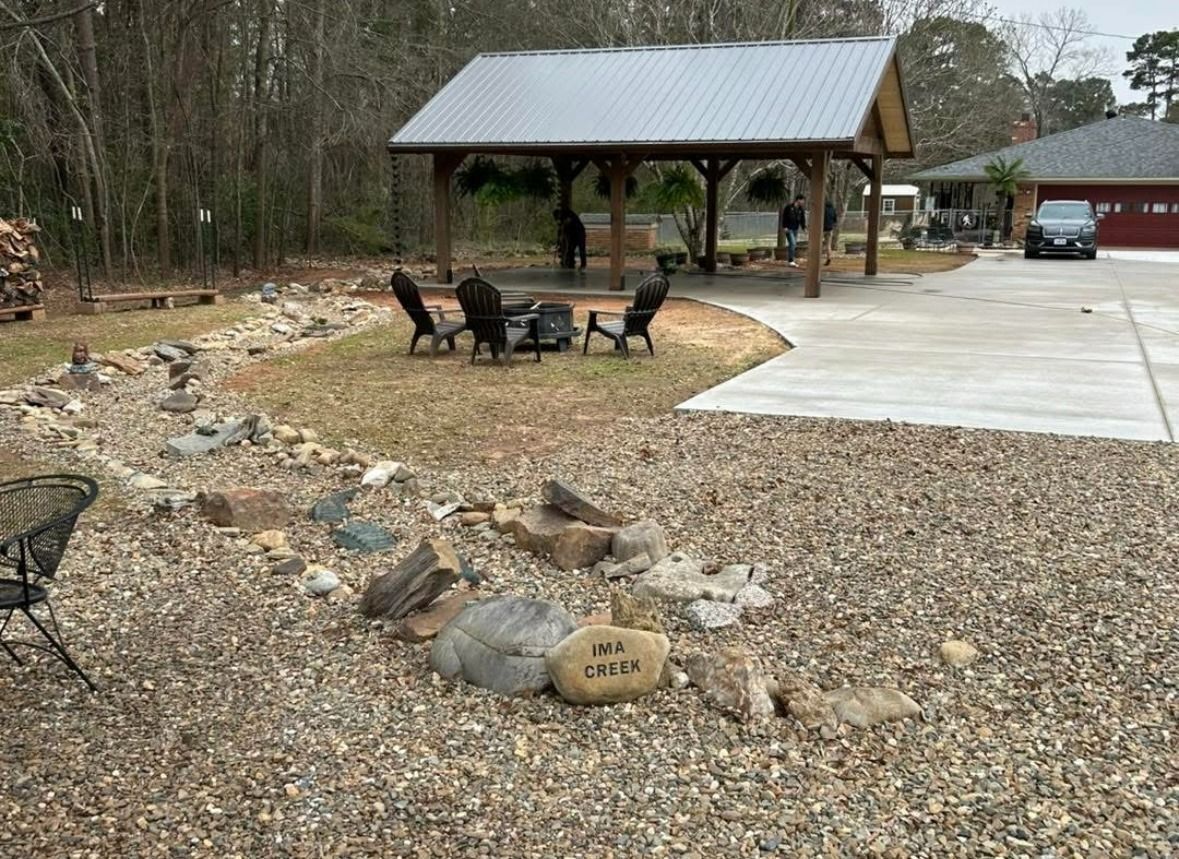 Gravel yard with fire pit, chairs, pavilion, and concrete driveway beside a red barn.