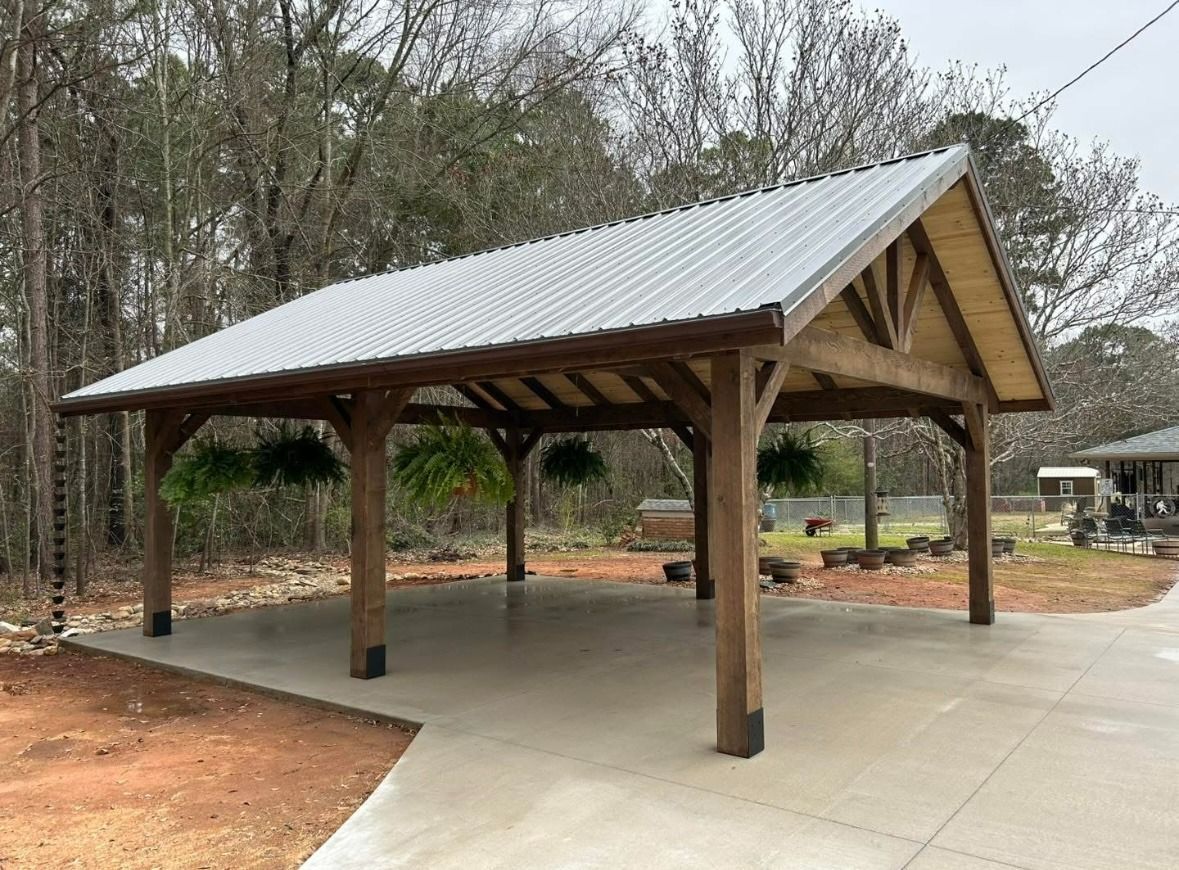 Open-sided wooden pavilion with metal roof on a concrete pad in a wooded park setting