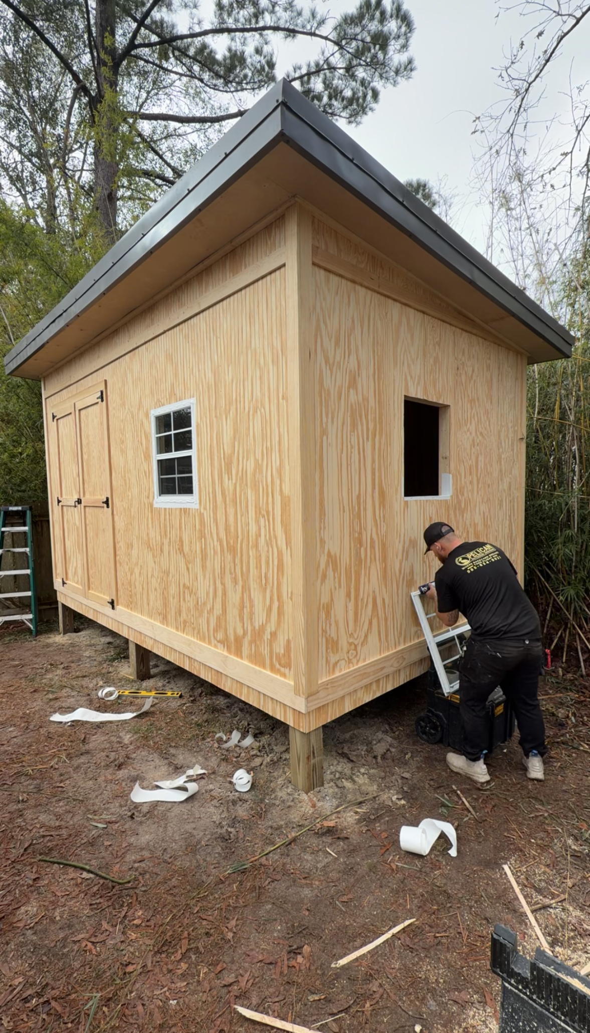 Small unfinished plywood shed with open window and door; person working beside it in a wooded yard.