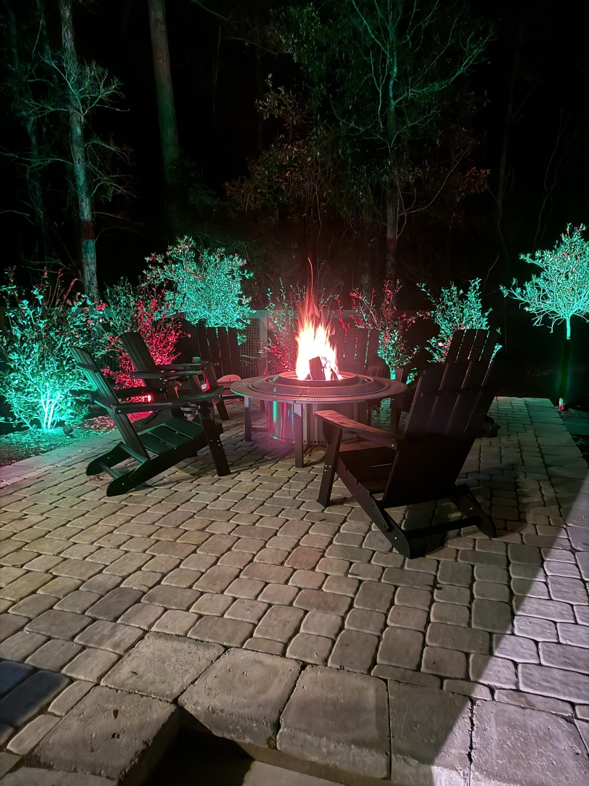 Firepit glowing on a cobblestone patio at night, with Adirondack chairs and lit trees around it