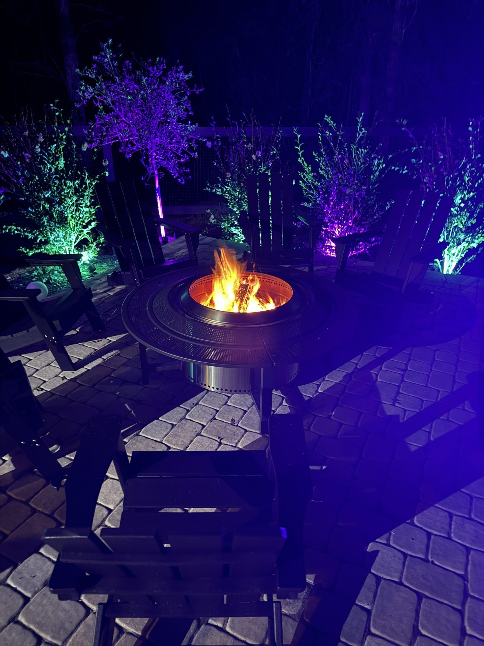 Outdoor patio fire pit at night with colorful purple and green lights illuminating chairs and trees.