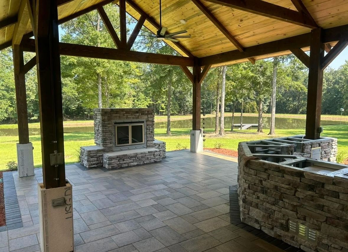 Covered outdoor pavilion with stone fire pits and paved floor in a grassy park.