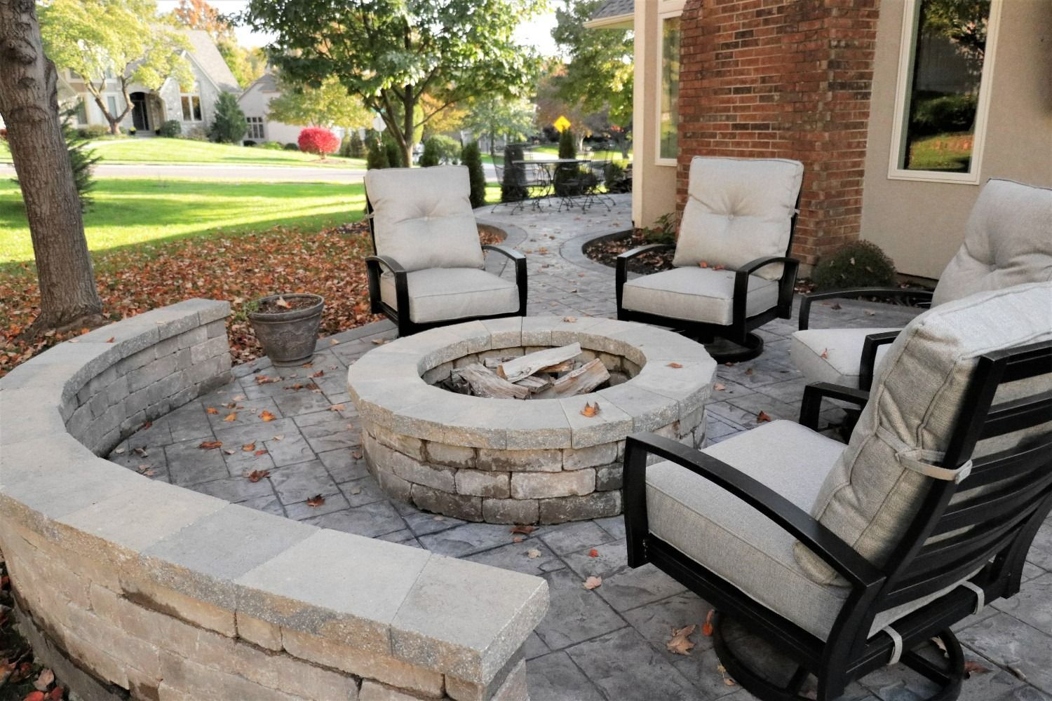 Stone patio with cushioned chairs arranged around a circular fire pit beside a brick house.