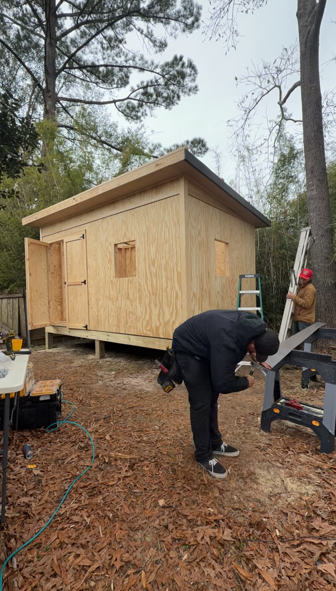 Two people assembling a small wooden shed in a wooded yard