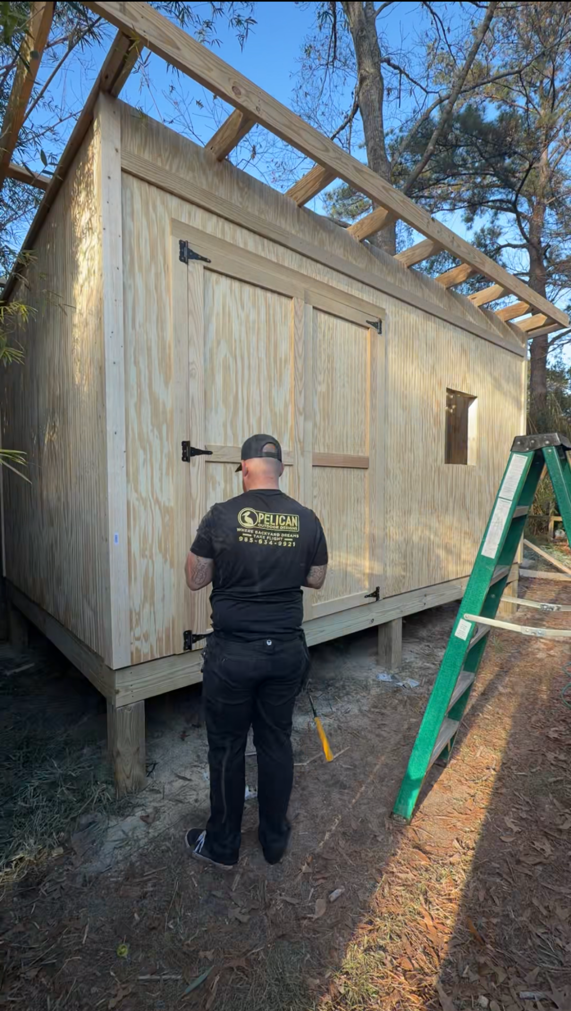 Worker painting exterior of a small wooden cabin beside a green ladder and trees