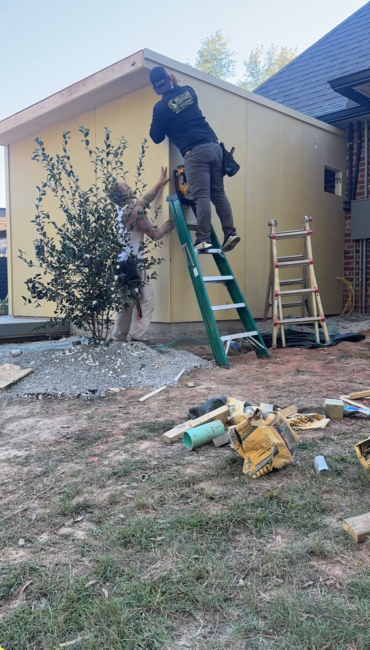 Person on a green ladder working on the roof edge of a beige house with tools in the yard.