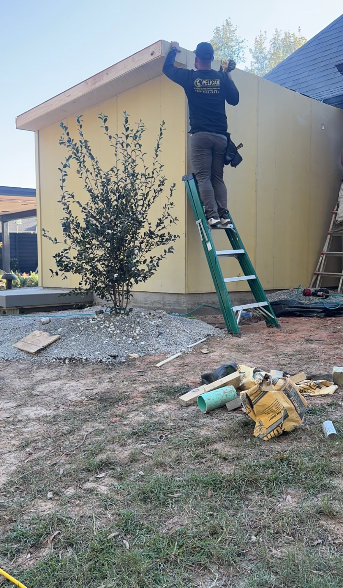 Person standing on a ladder working on the roofline of a beige house with tools on the ground.