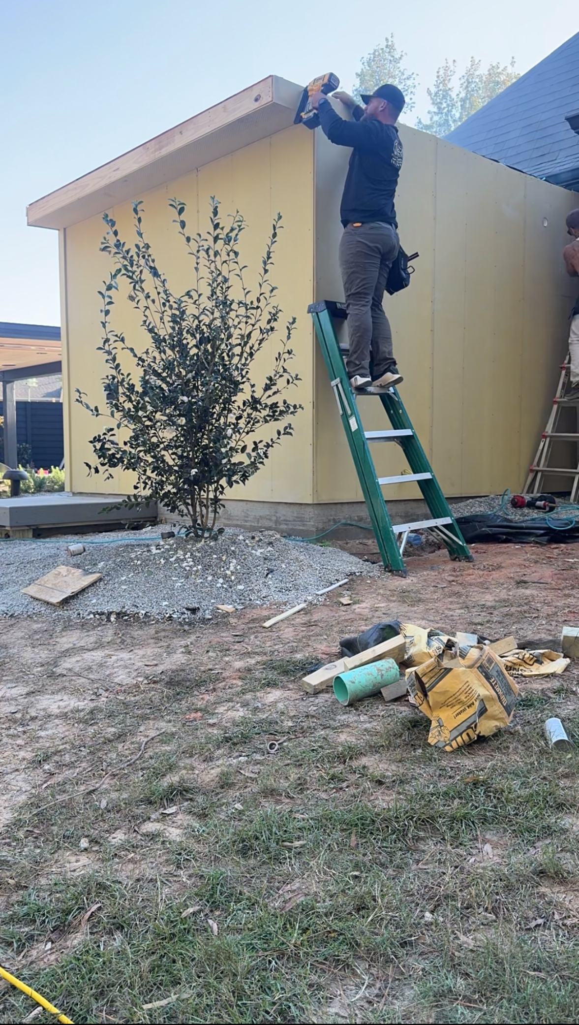 Worker on green ladder repairing the roof edge of a yellow shed beside a small tree.