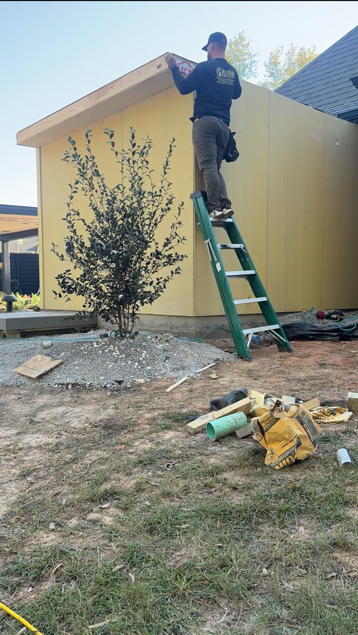 Worker on a green ladder painting the upper wall of a yellow house outdoors.