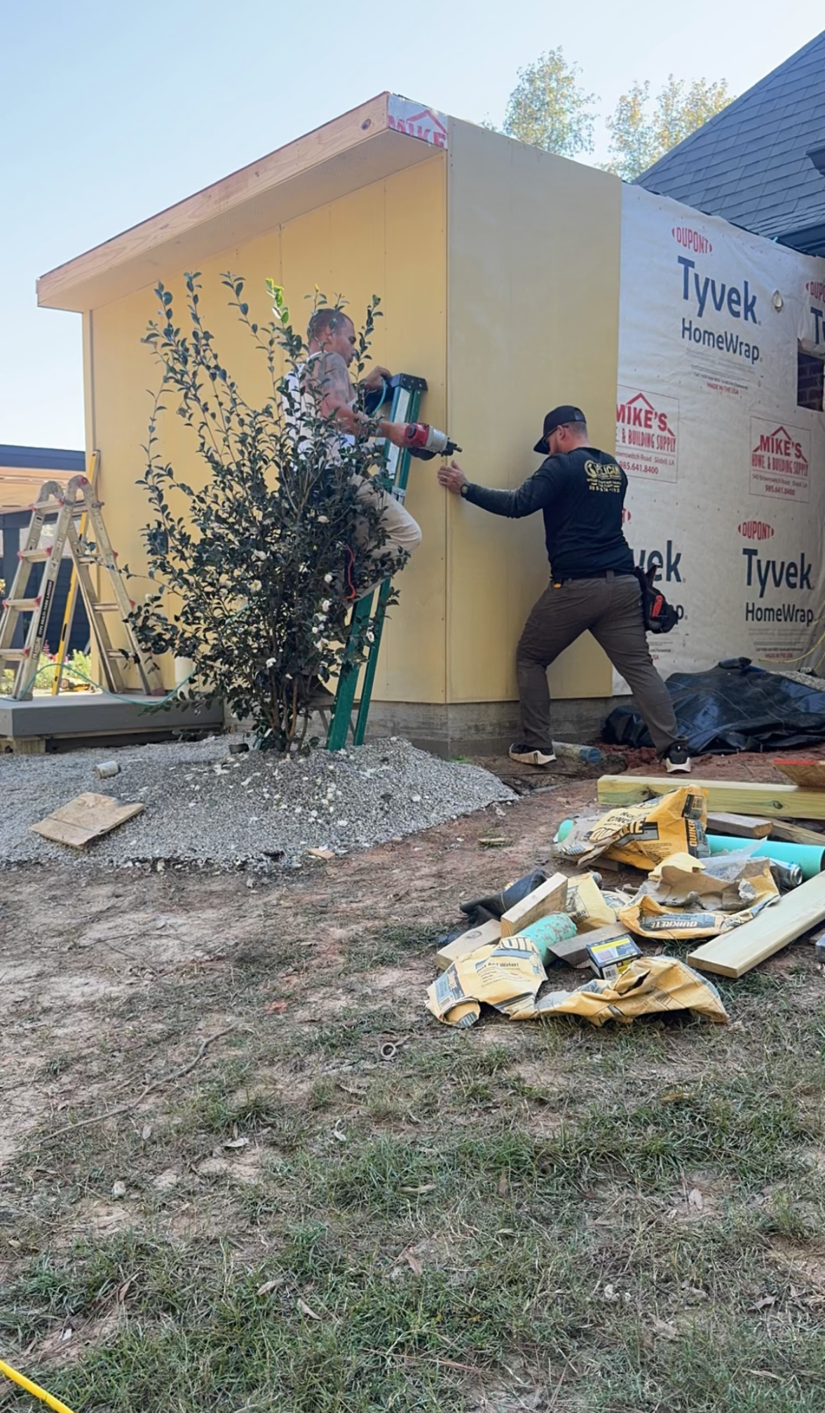 Two workers installing siding on a small yellow house, with construction debris in the yard.