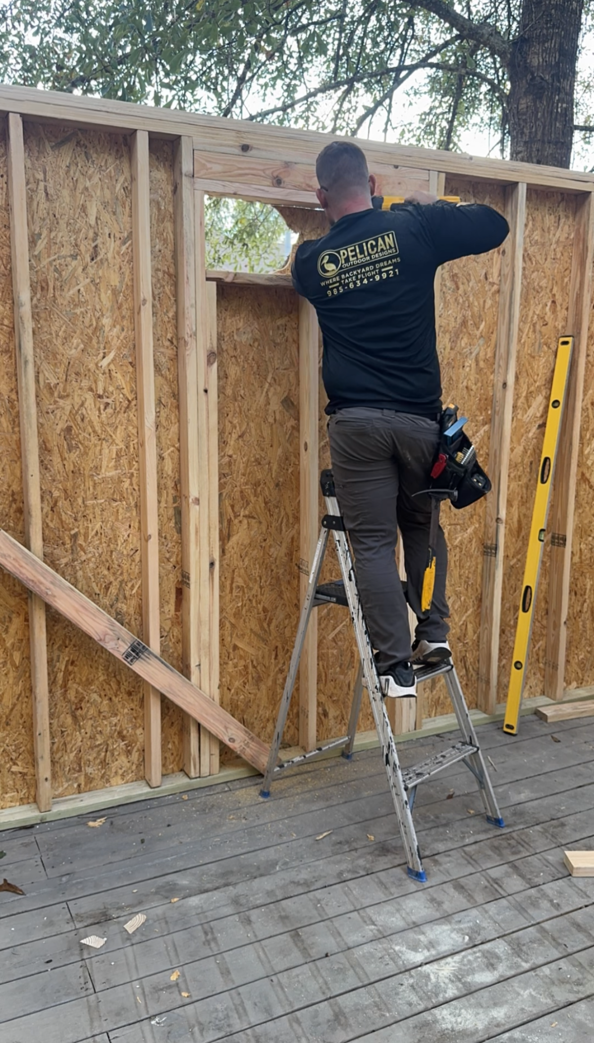 Worker standing on a ladder fastening plywood sheathing to a framed wall outdoors