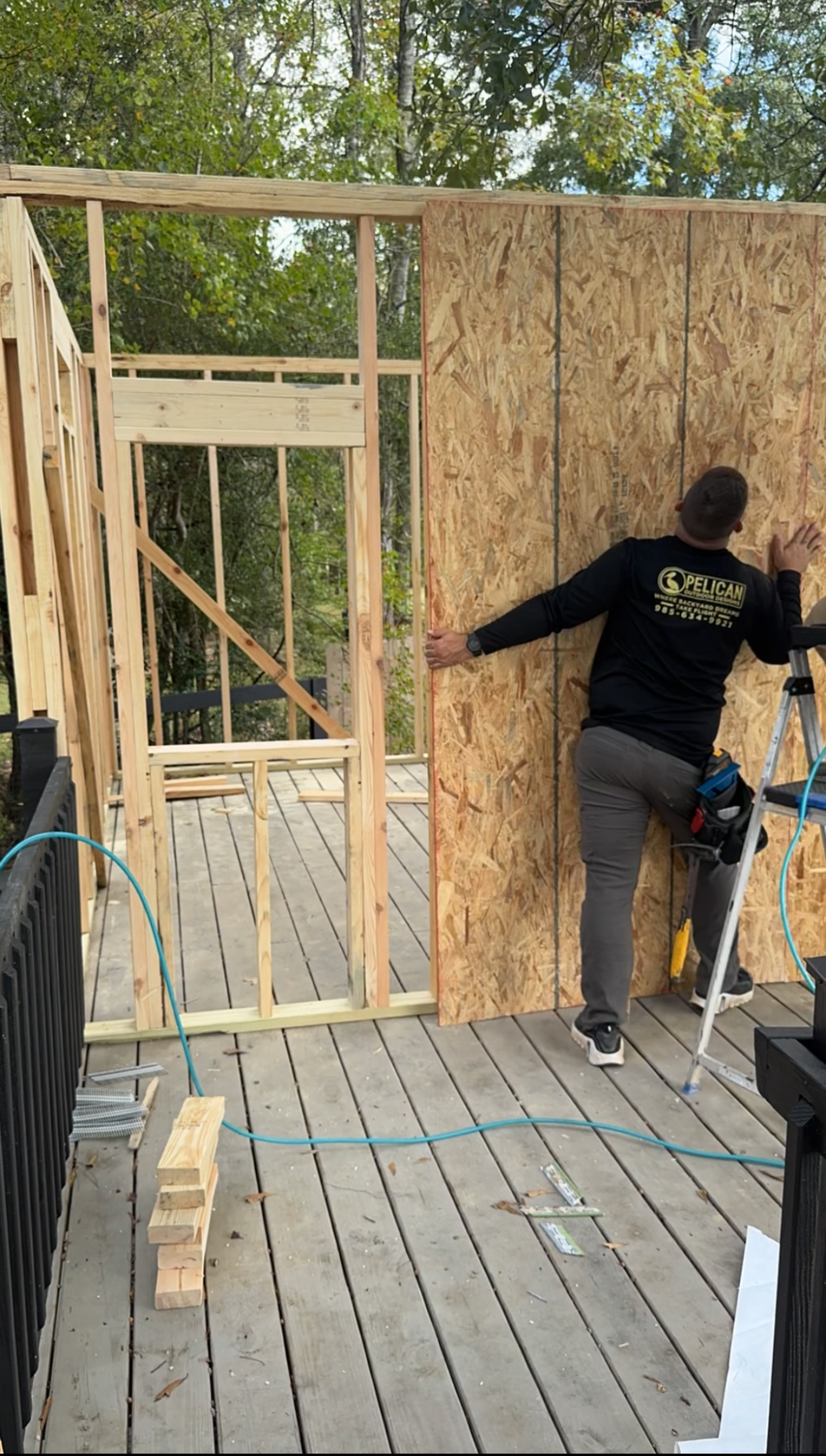 Person working on a wood-framed wall on an outdoor deck, holding a tool next to plywood sheathing.