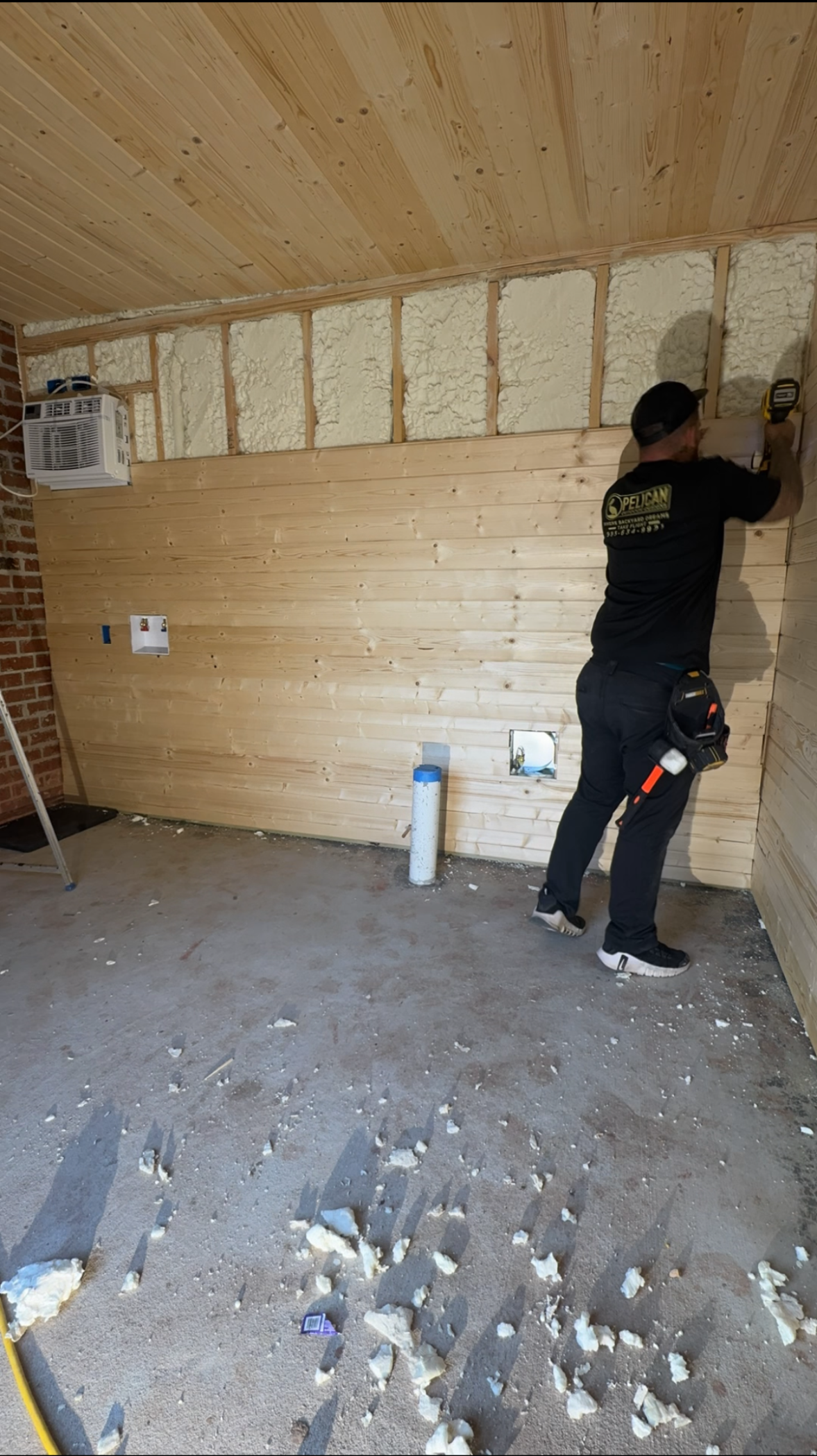 Person installing insulation on a wood-paneled wall inside an unfinished room.