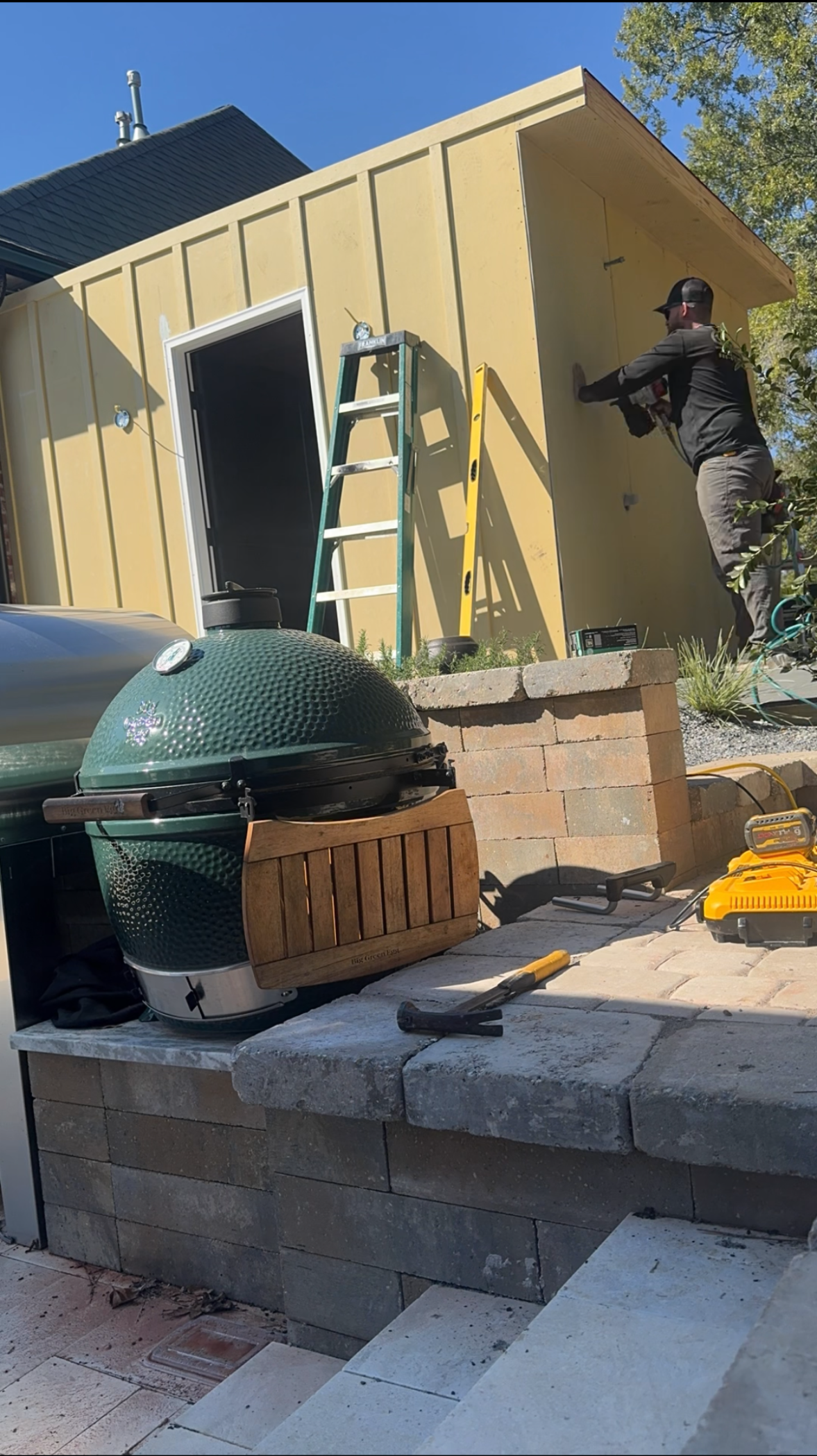 Person working beside a small yellow shed with an open door, tools, and a green dome grill in front
