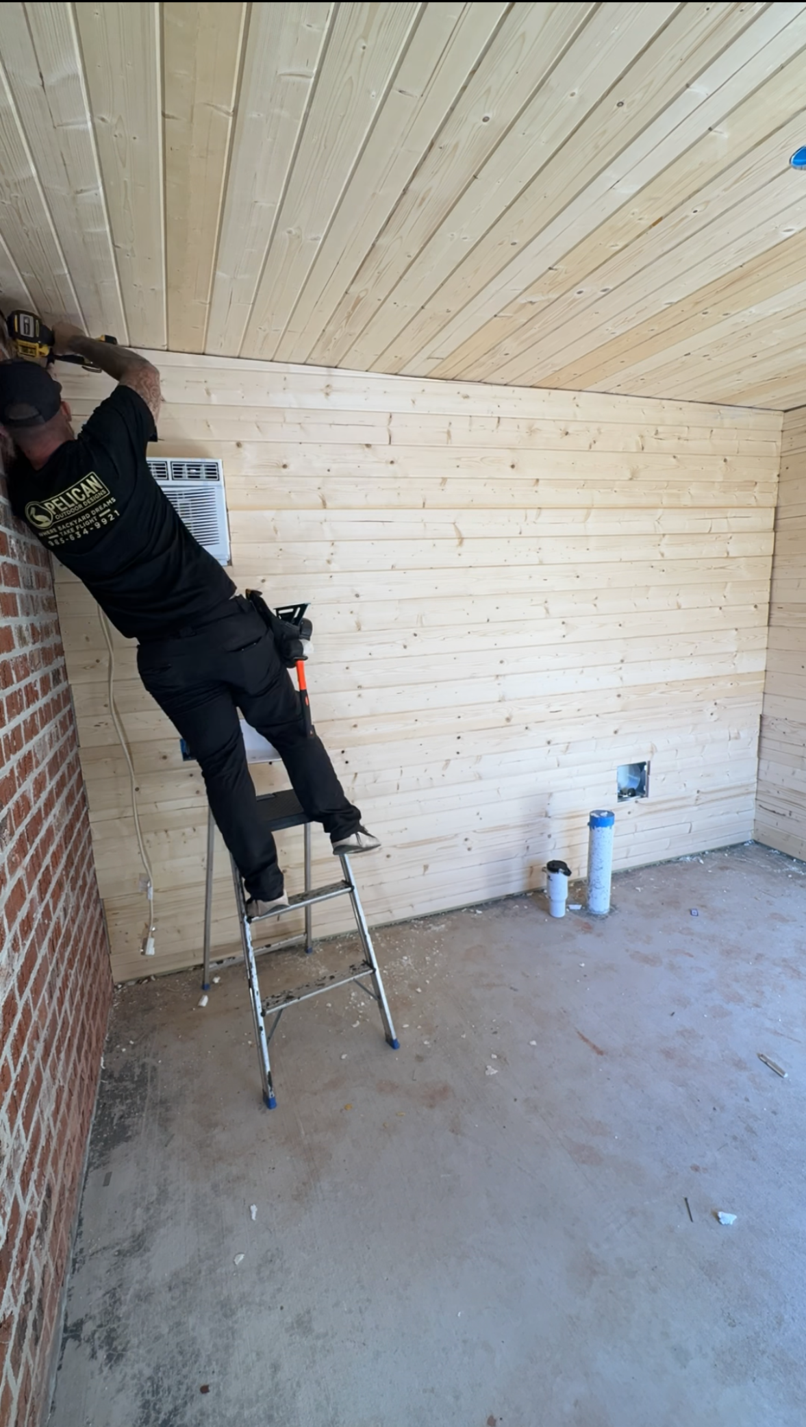 Person on a ladder painting a light wood-paneled ceiling in an unfinished room.