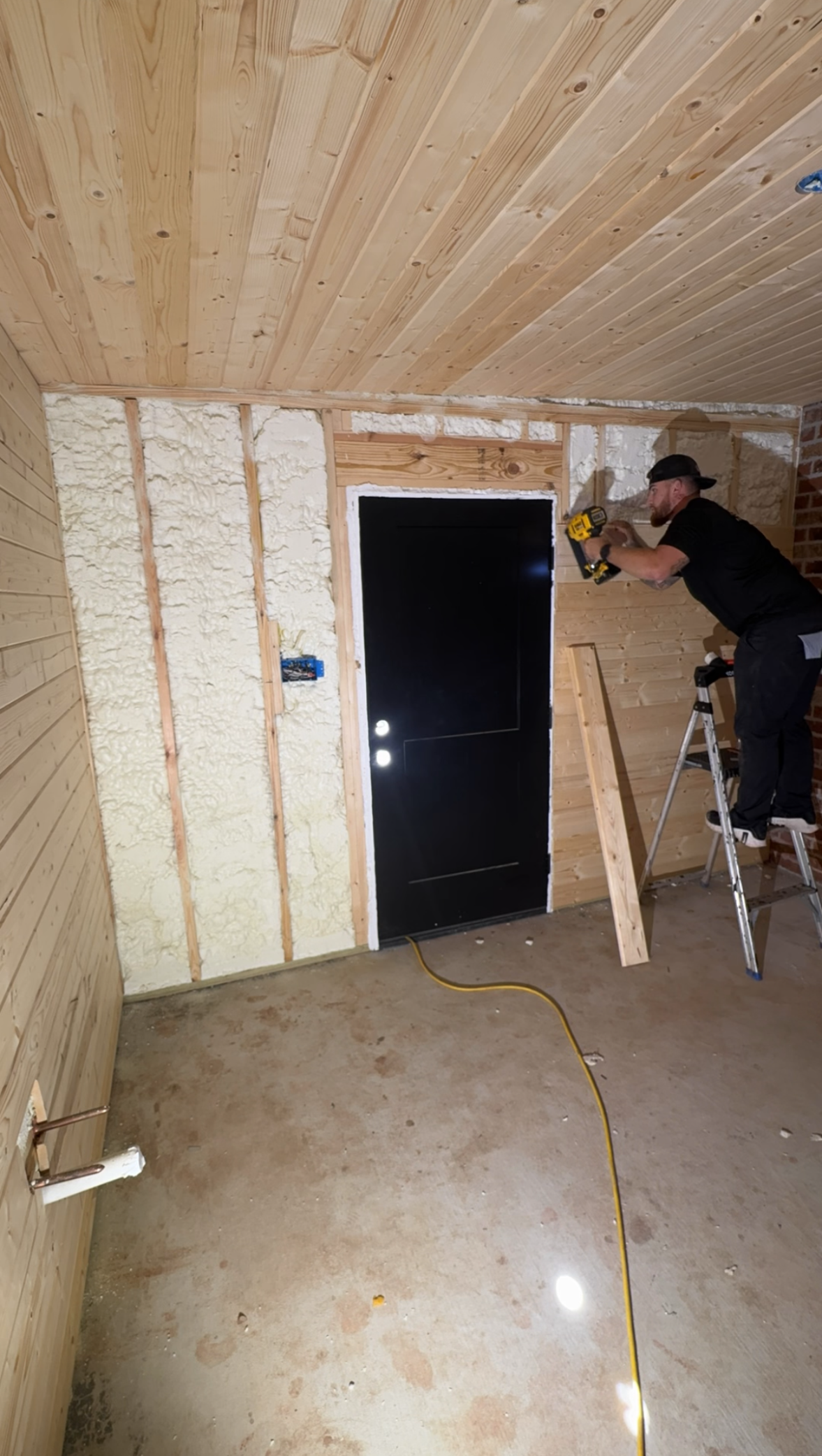 Worker installing insulation in an unfinished room with exposed foam walls and a black door.