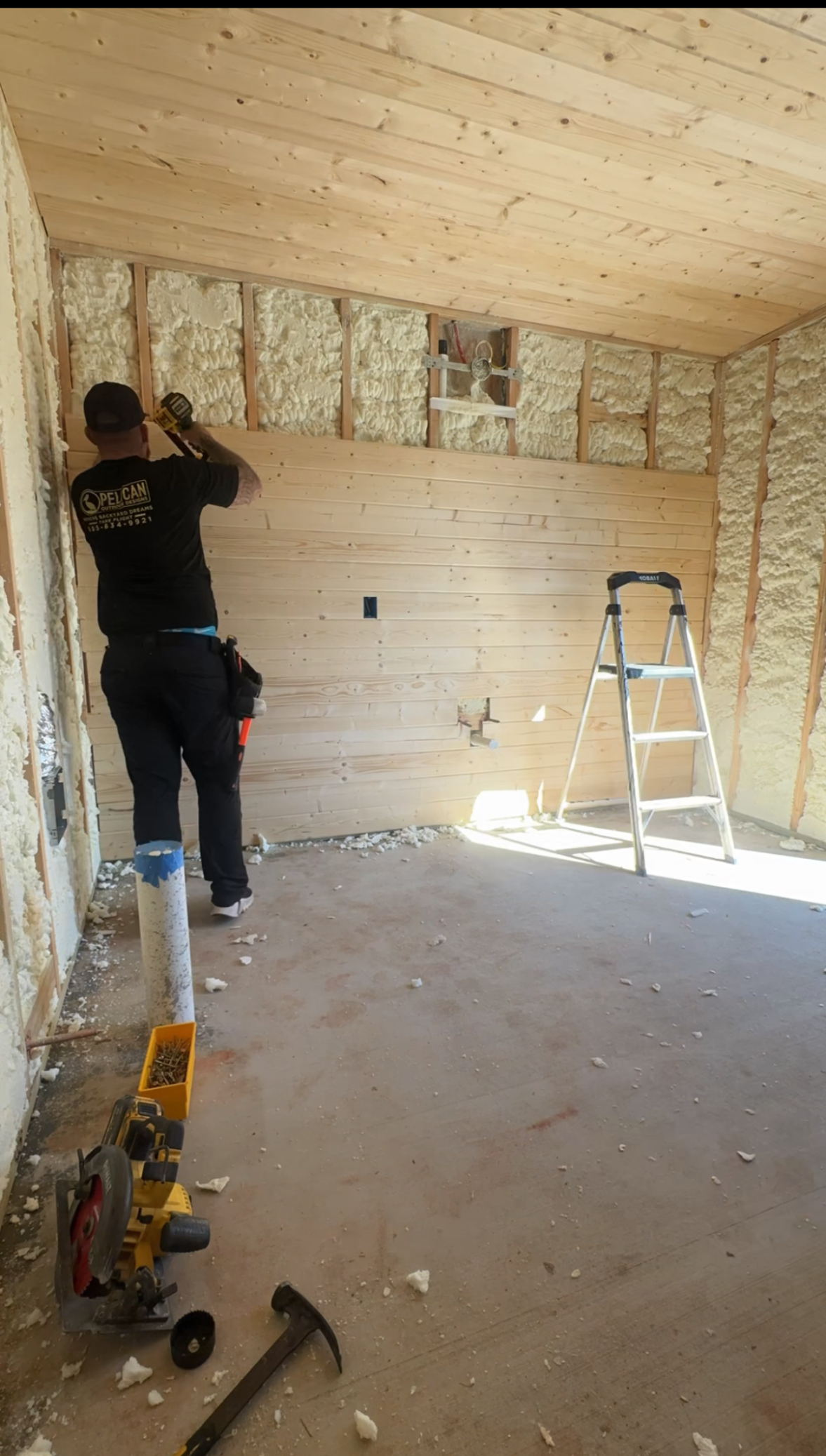 Person working on a wall inside an unfinished room with tools and a ladder nearby.