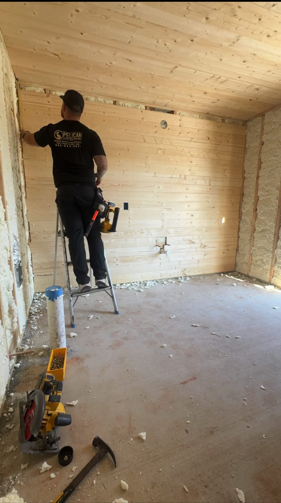 Worker on a ladder nailing light wood paneling to a wall in a bare room
