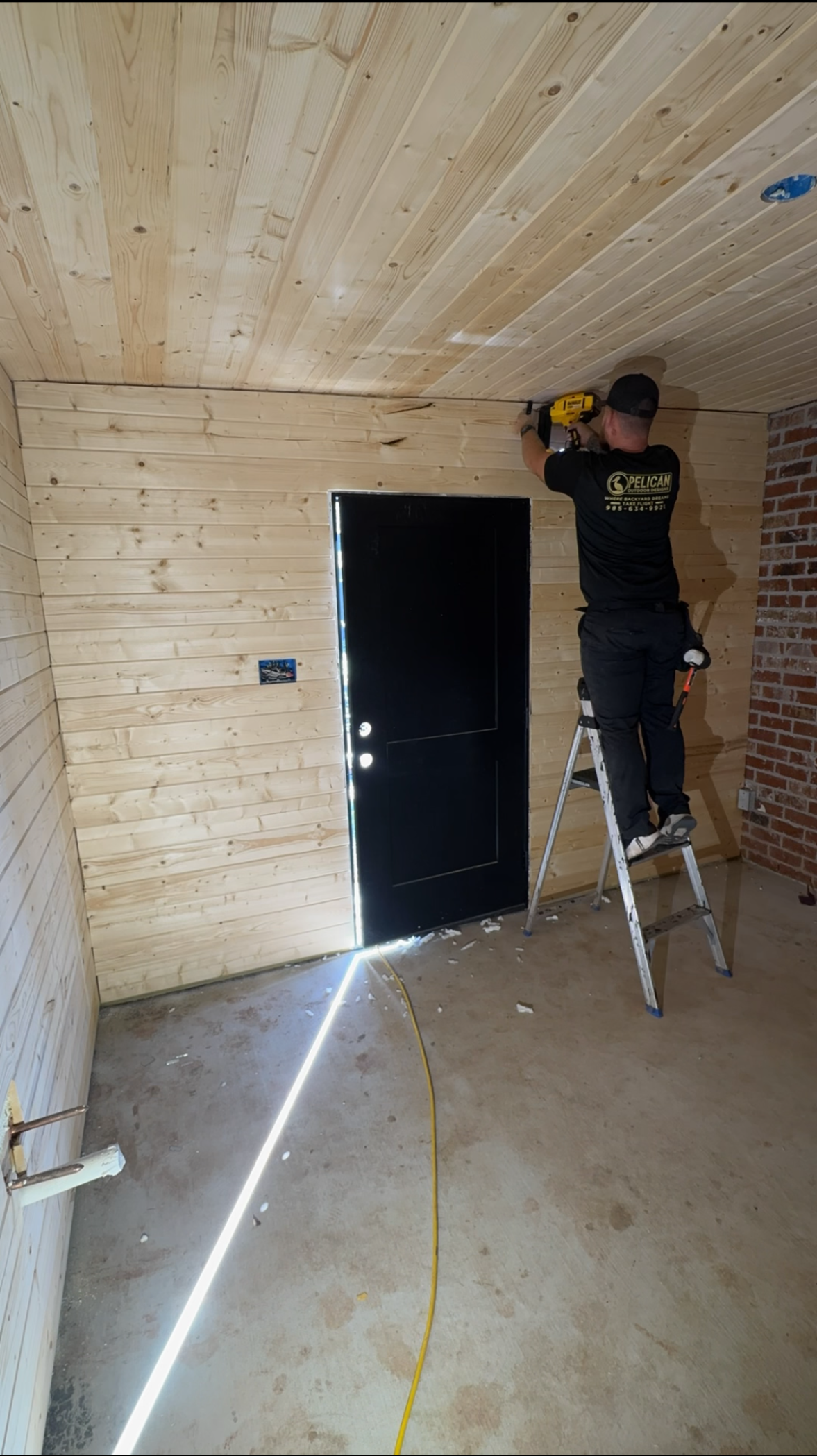 Worker installing a wooden ceiling in a narrow unfinished room with a black door and ladder.