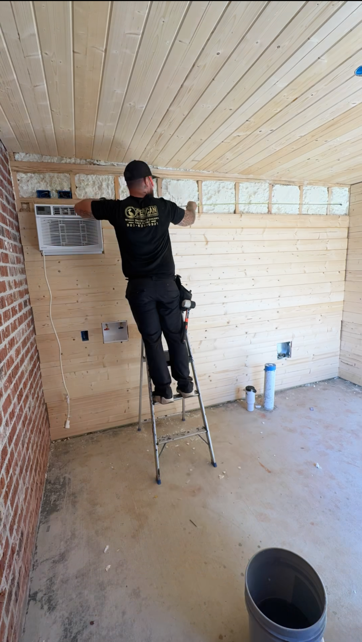 Person standing on a small ladder, installing wood paneling on a wall in a room under construction.