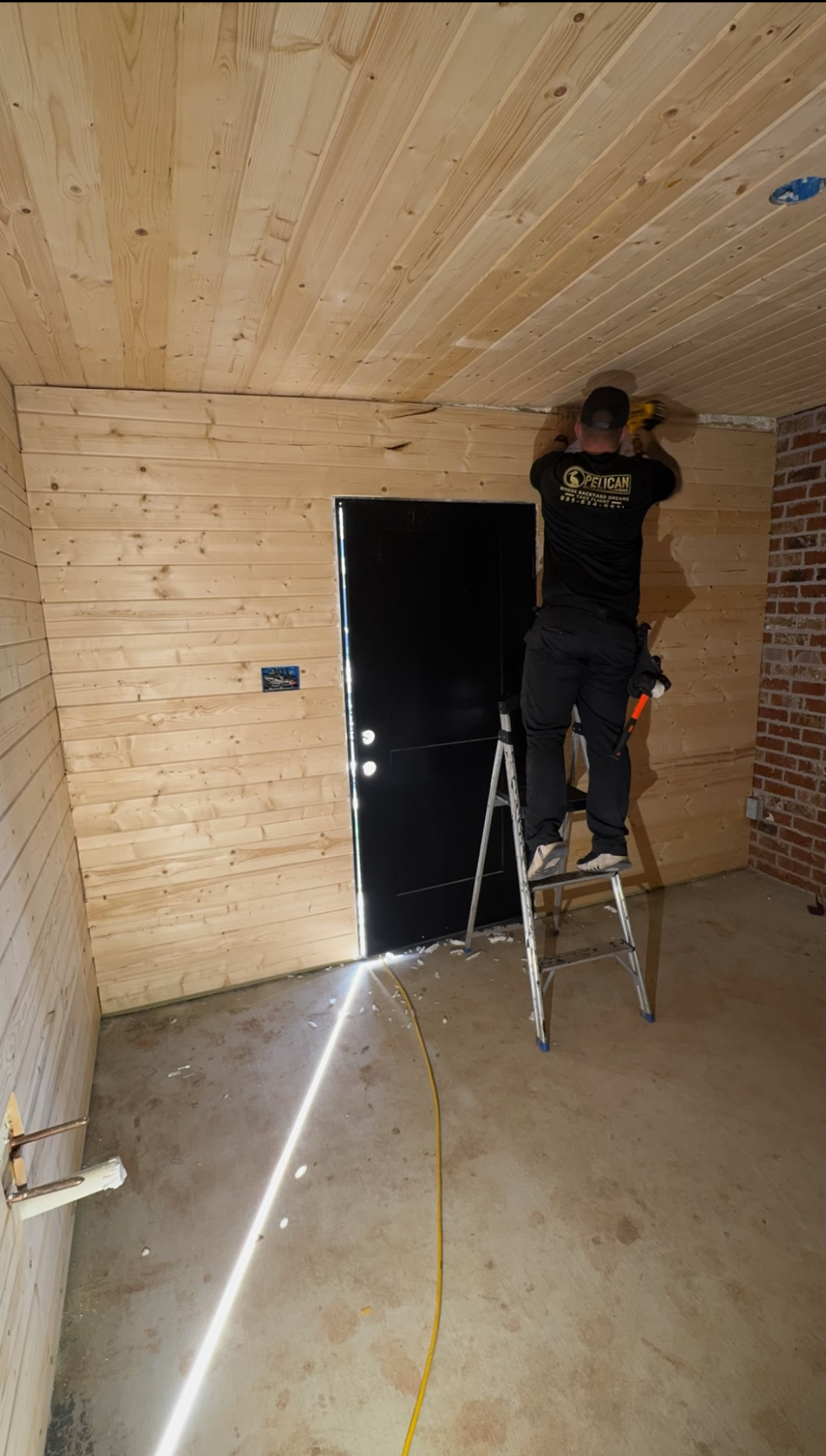 Person on a ladder working on a wood-paneled room interior beside a black door.