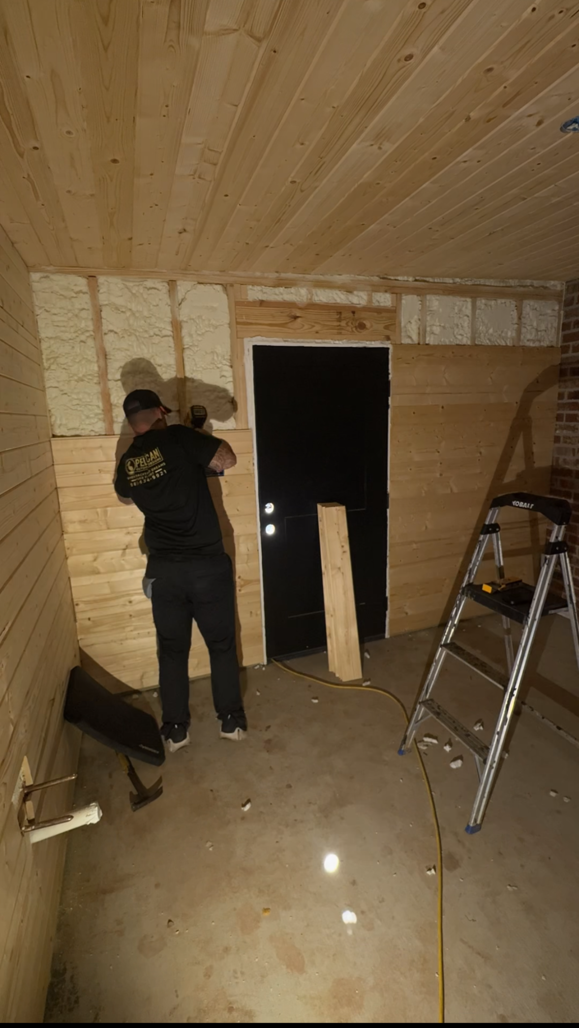 Worker installing wall panels in a wood-paneled room with a ladder and tools on the floor.