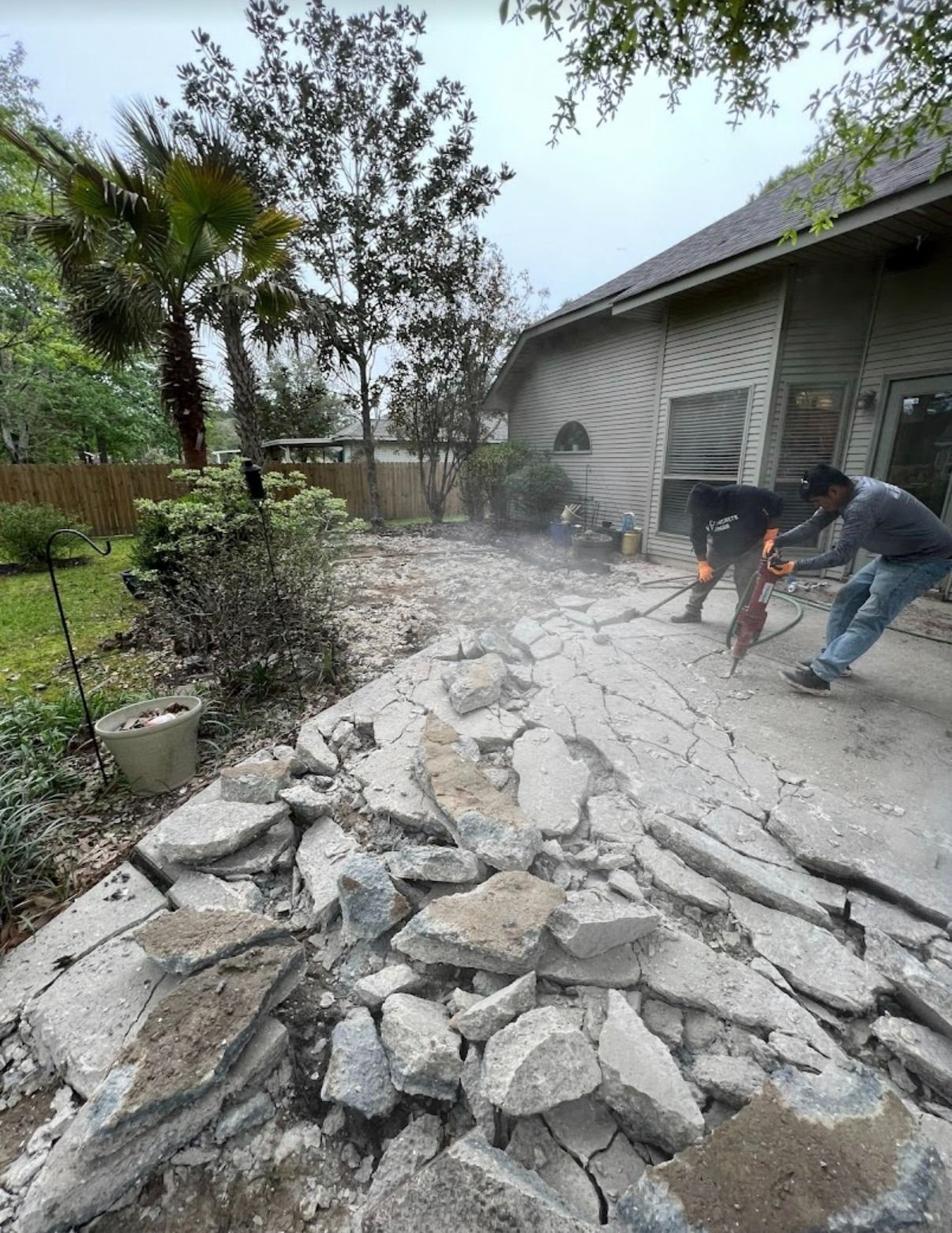 Workers jackhammering a broken concrete patio beside a house, with rubble piled in the foreground.