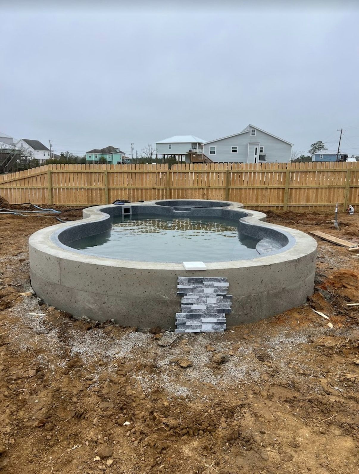 Round concrete hot tub under construction in a dirt yard with a wooden fence and houses in the background