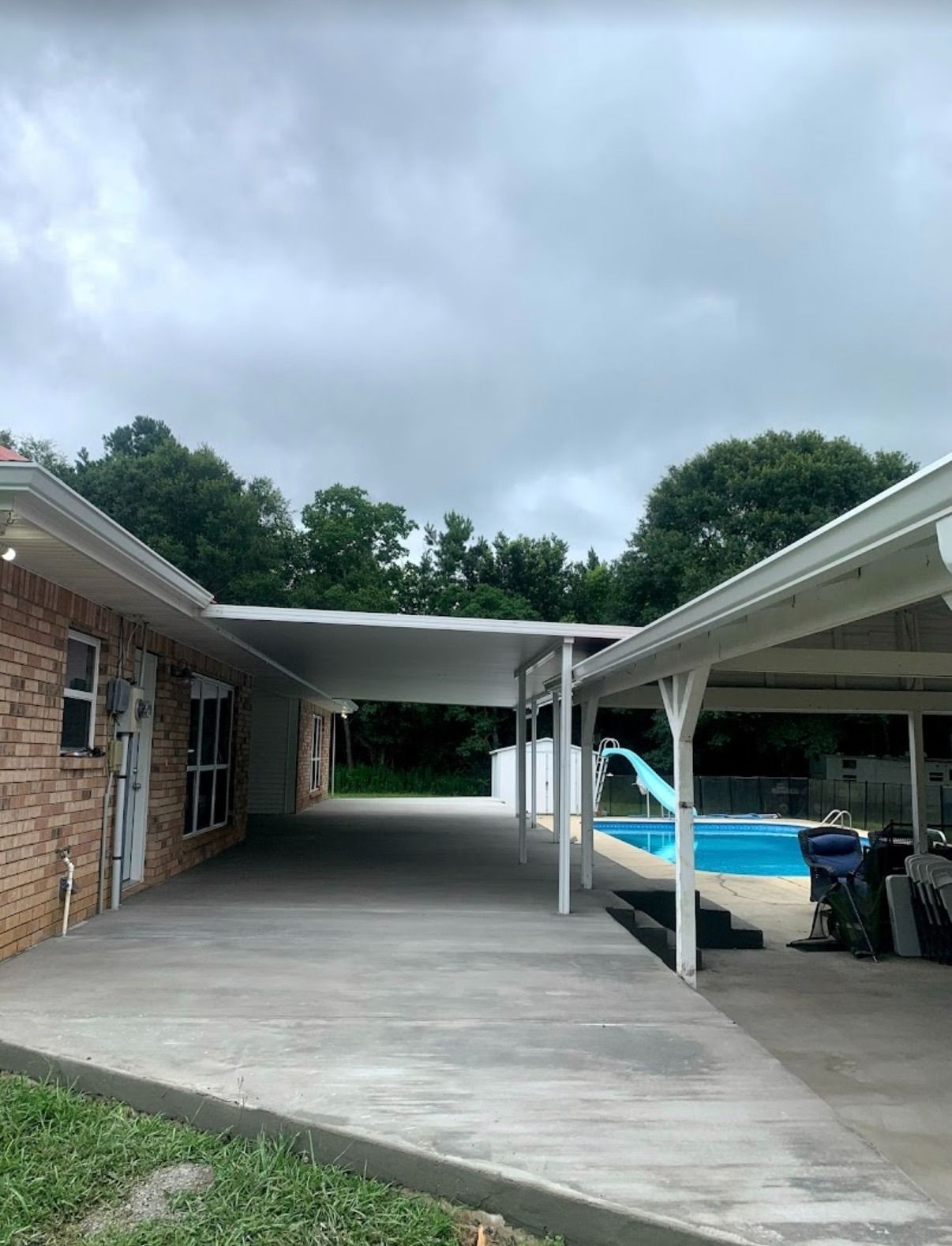 Covered driveway beside a brick house, with a white canopy and a blue pool in the background.