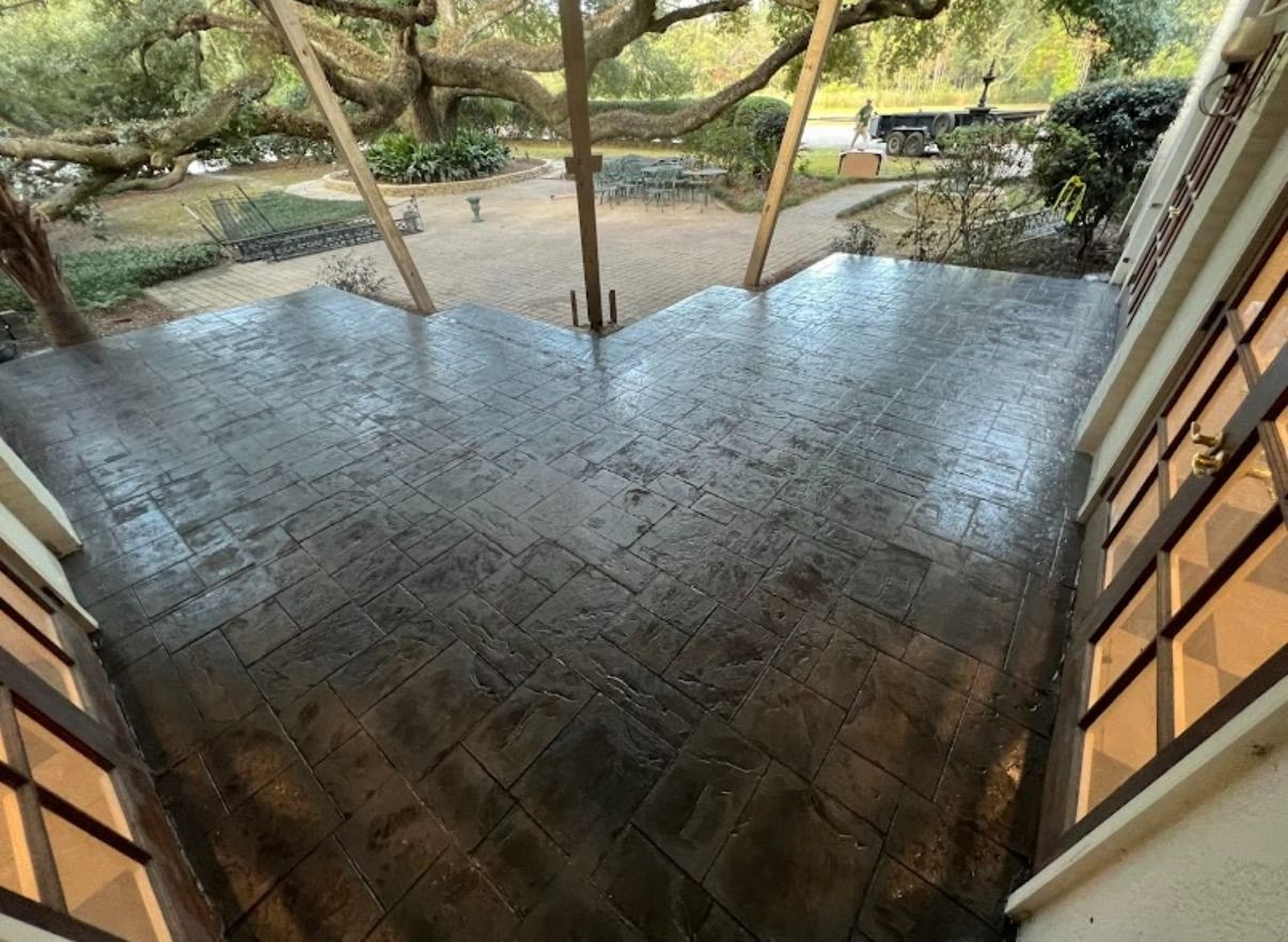 Shiny dark brick patio under trees beside a building, with a wet surface and a pathway beyond.