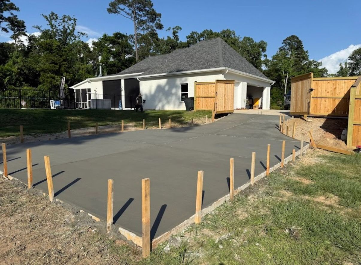 Freshly poured concrete driveway at a house, bordered by wooden stakes and forms, with trees and fences nearby