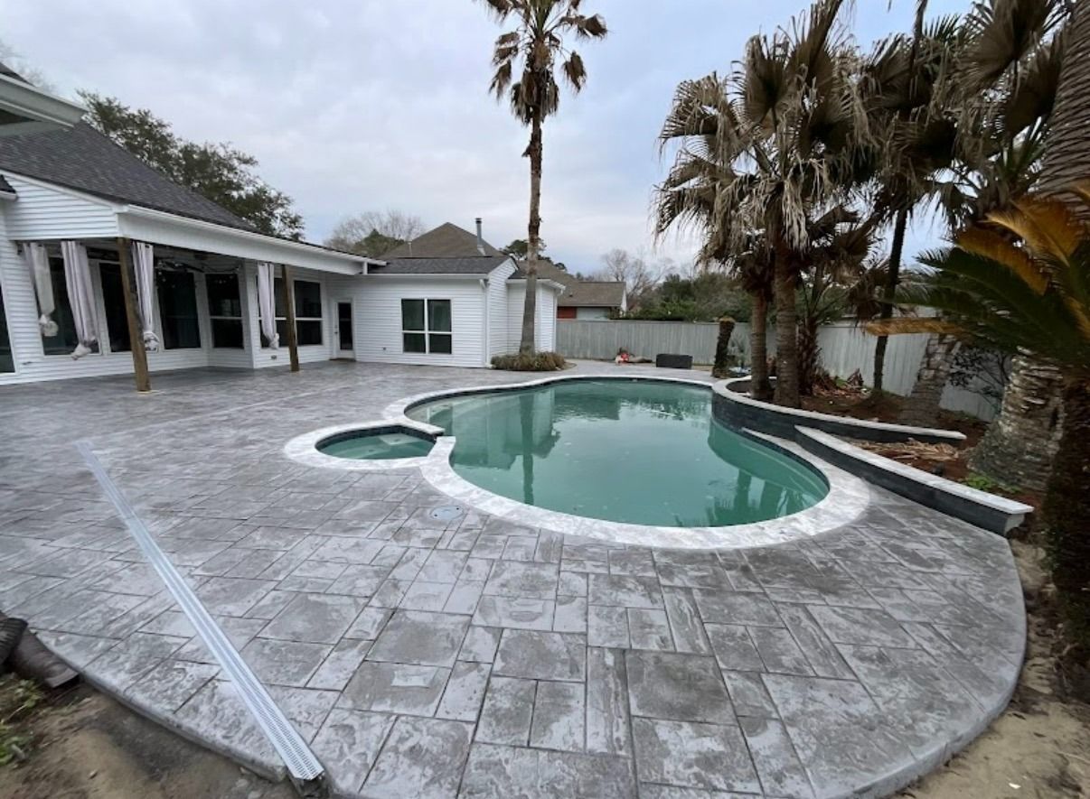Patio with a curved swimming pool beside a house, surrounded by palm trees and gray stone paving