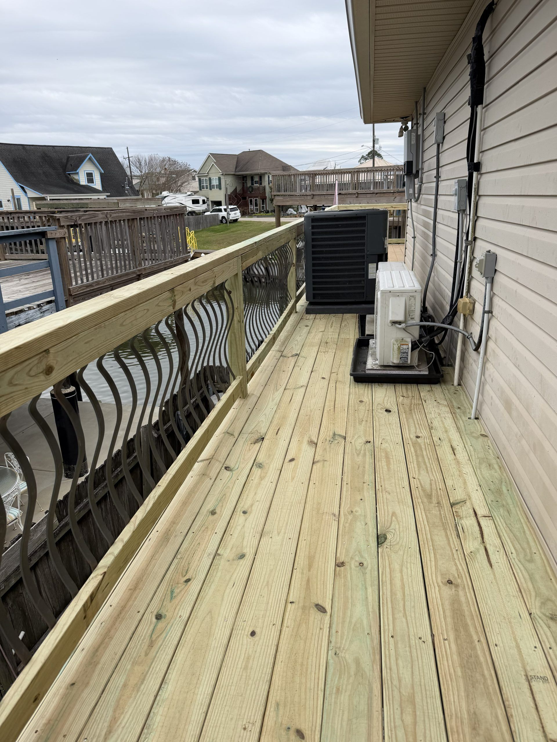 Wooden deck beside a house, with railing, air conditioner units, and a cloudy suburban neighborhood view.
