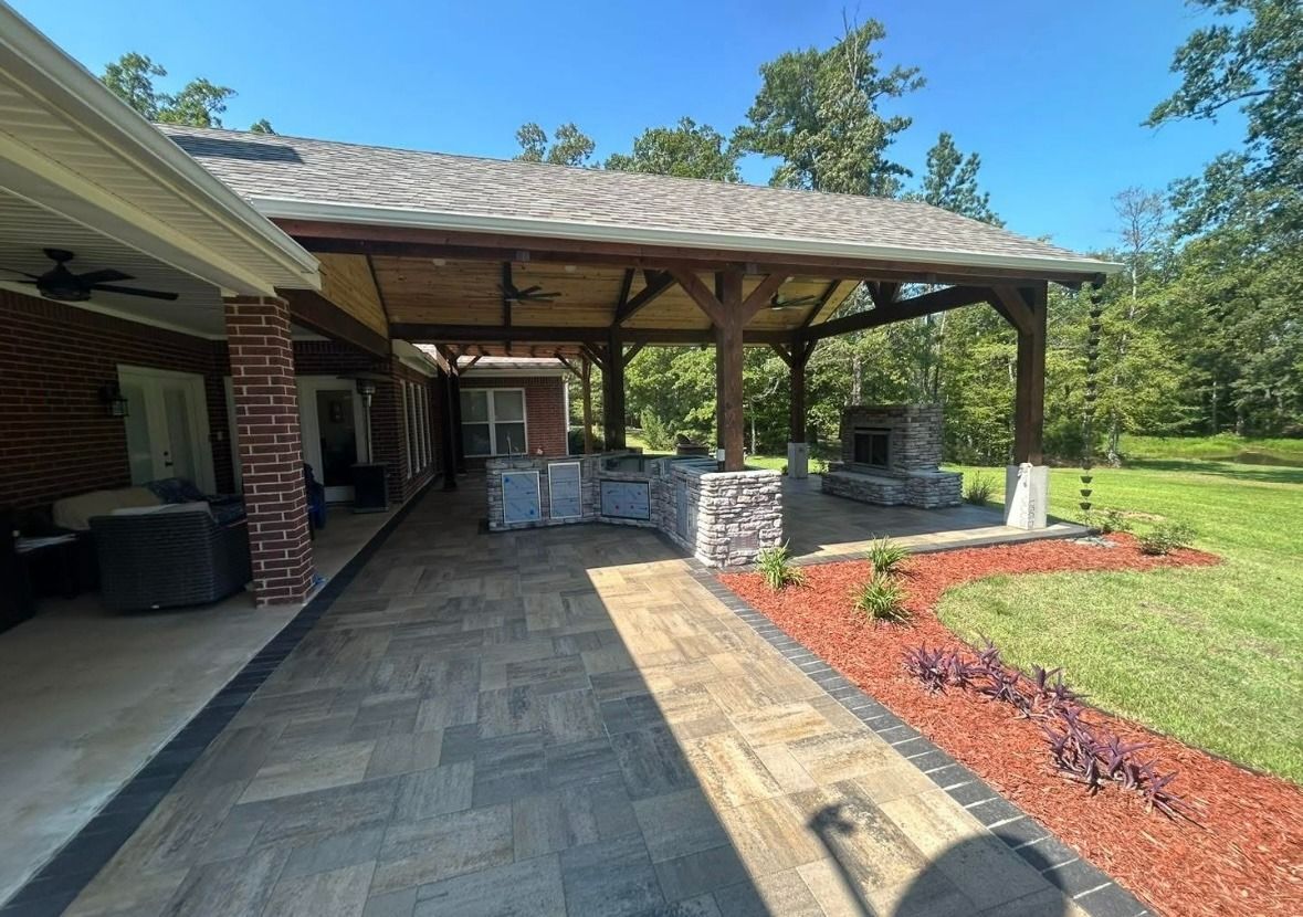 Covered stone patio with outdoor kitchen, brick columns, and landscaped lawn under a blue sky