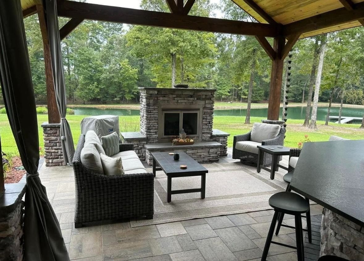 Covered patio seating area with wicker chairs around a stone fireplace and table, overlooking a grassy yard