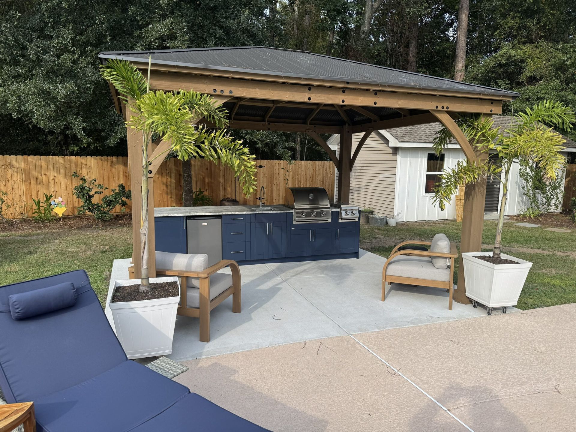 Covered backyard patio with blue seating, outdoor kitchen, and white planters under a wooden pergola
