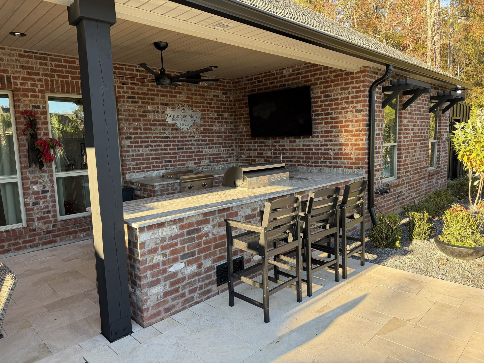 Covered brick patio with bar seating, wall-mounted TV, ceiling fan, and concrete floor