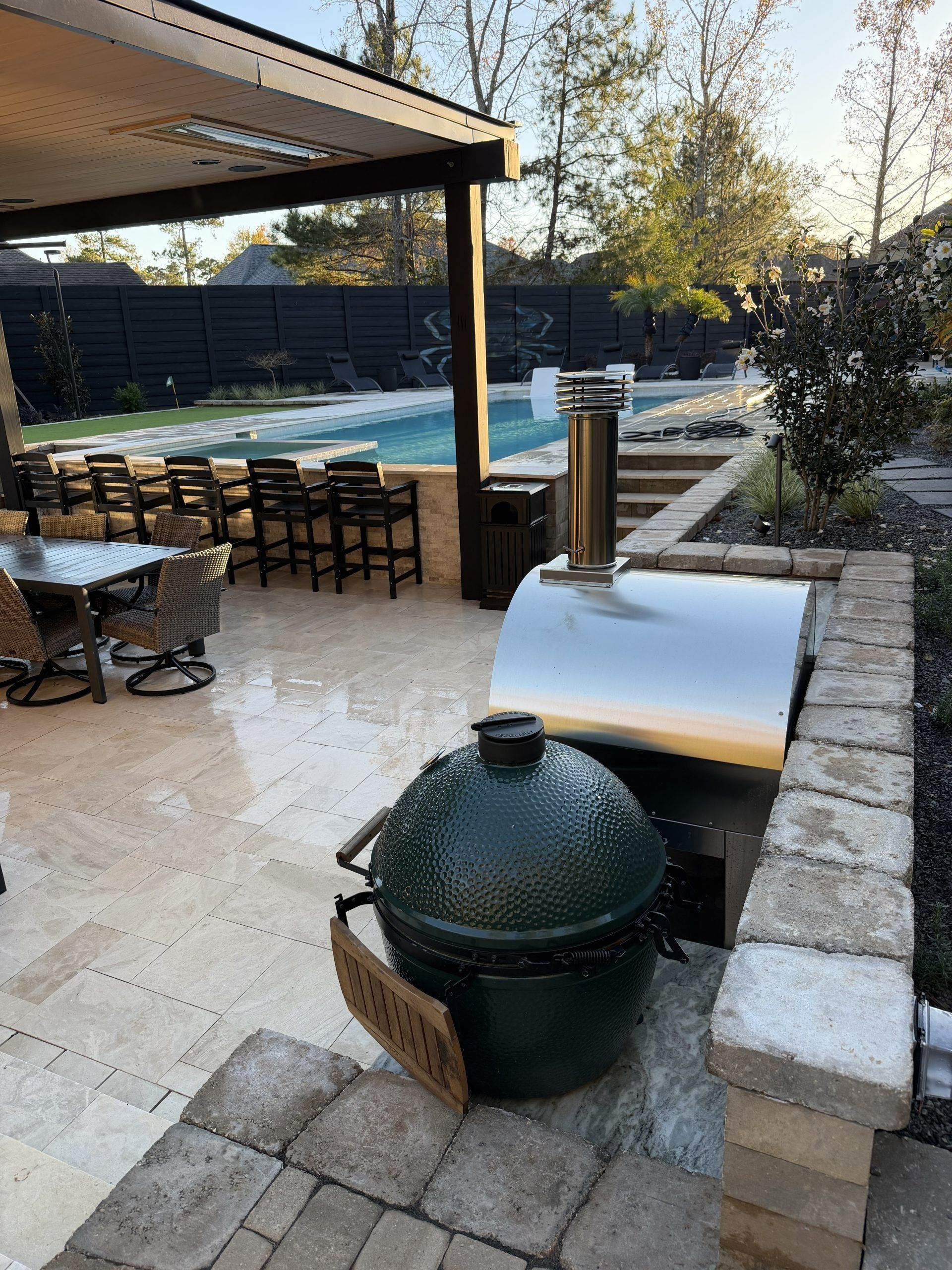 Backyard patio with a green kamado grill, stone seating wall, and swimming pool under a covered pergola.