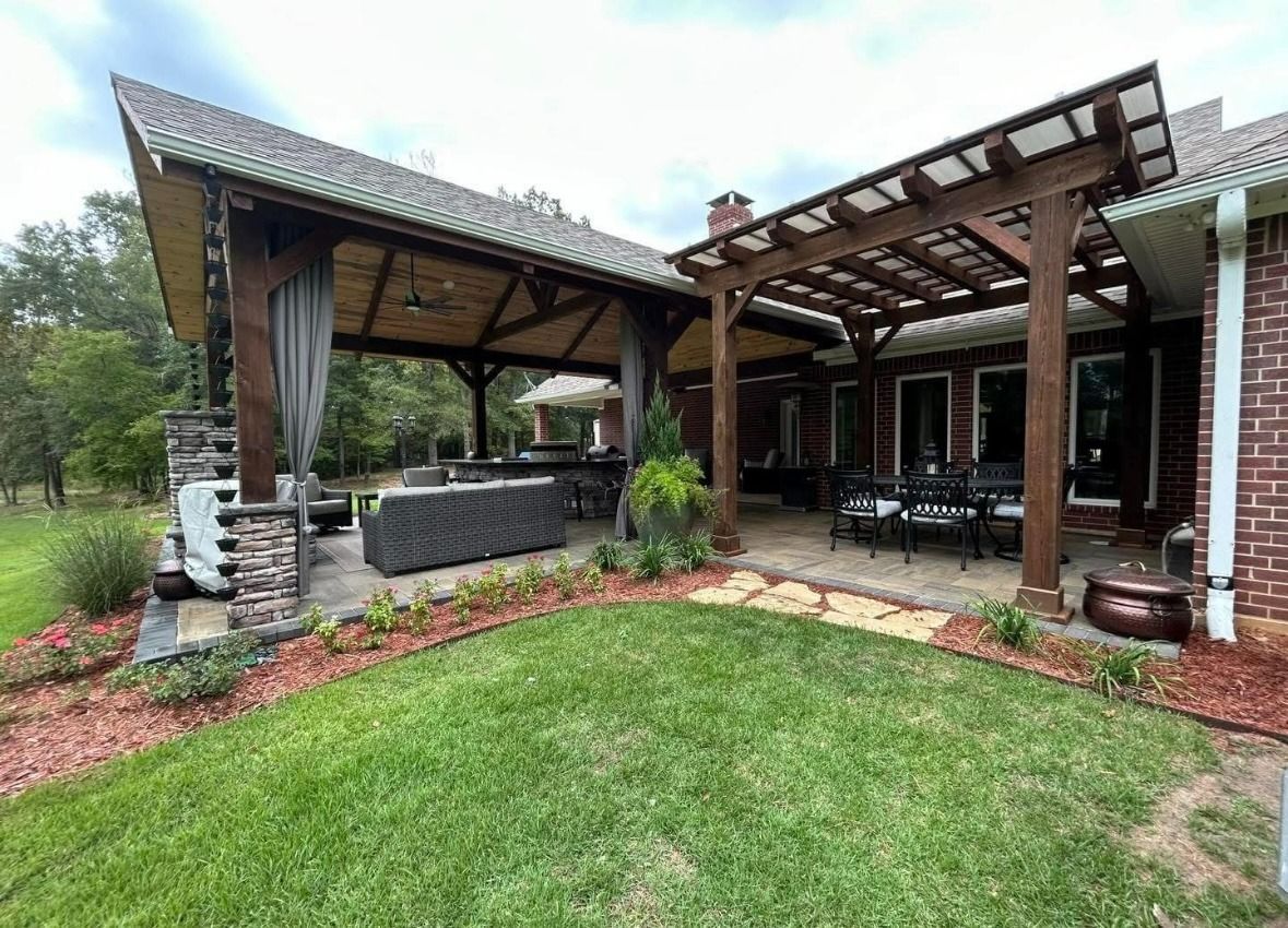 Covered patio with wooden pergola, outdoor seating, stone grill, and green lawn beside a brick house.