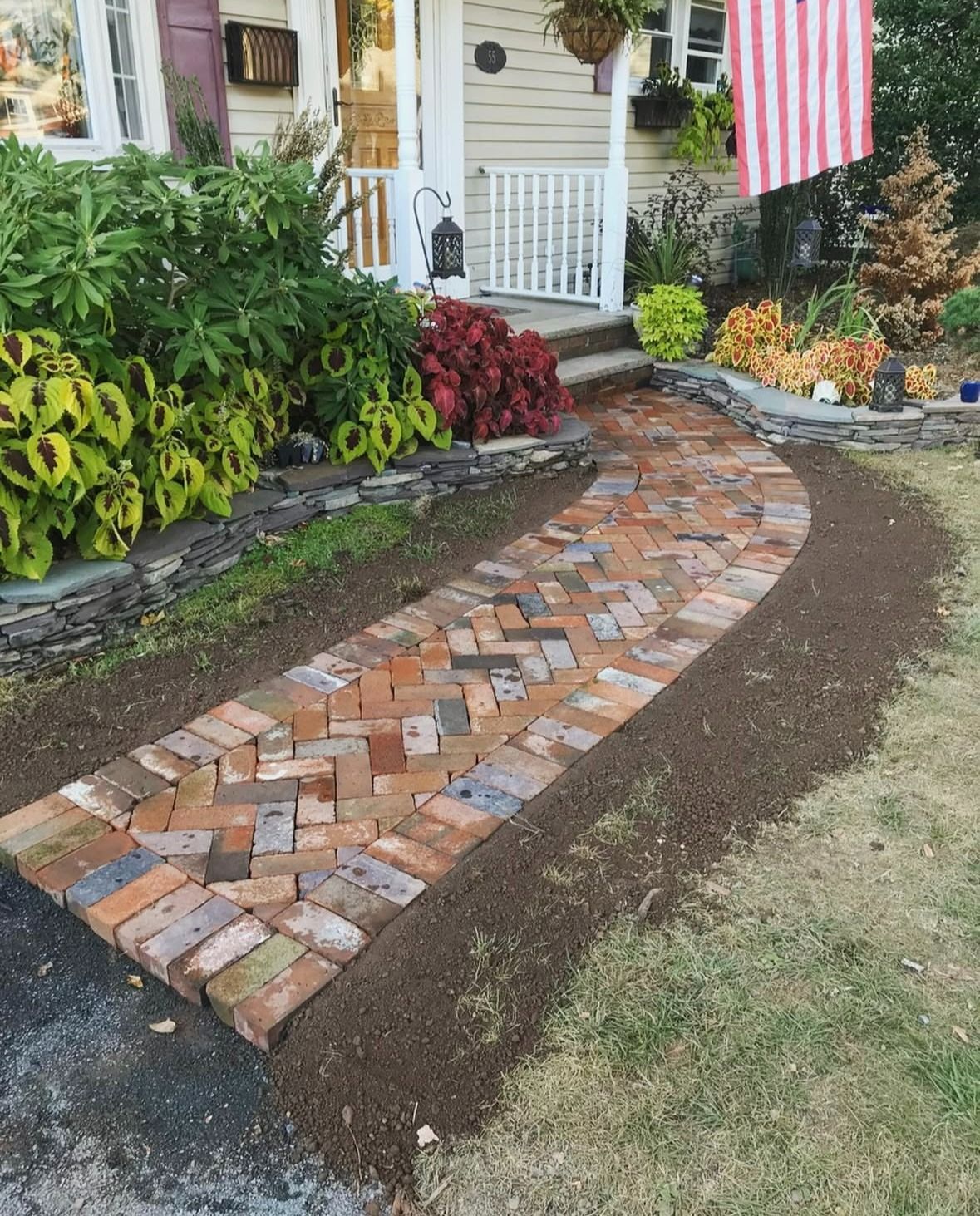 Curved brick garden walkway bordered by mulch and plants beside a house with an American flag